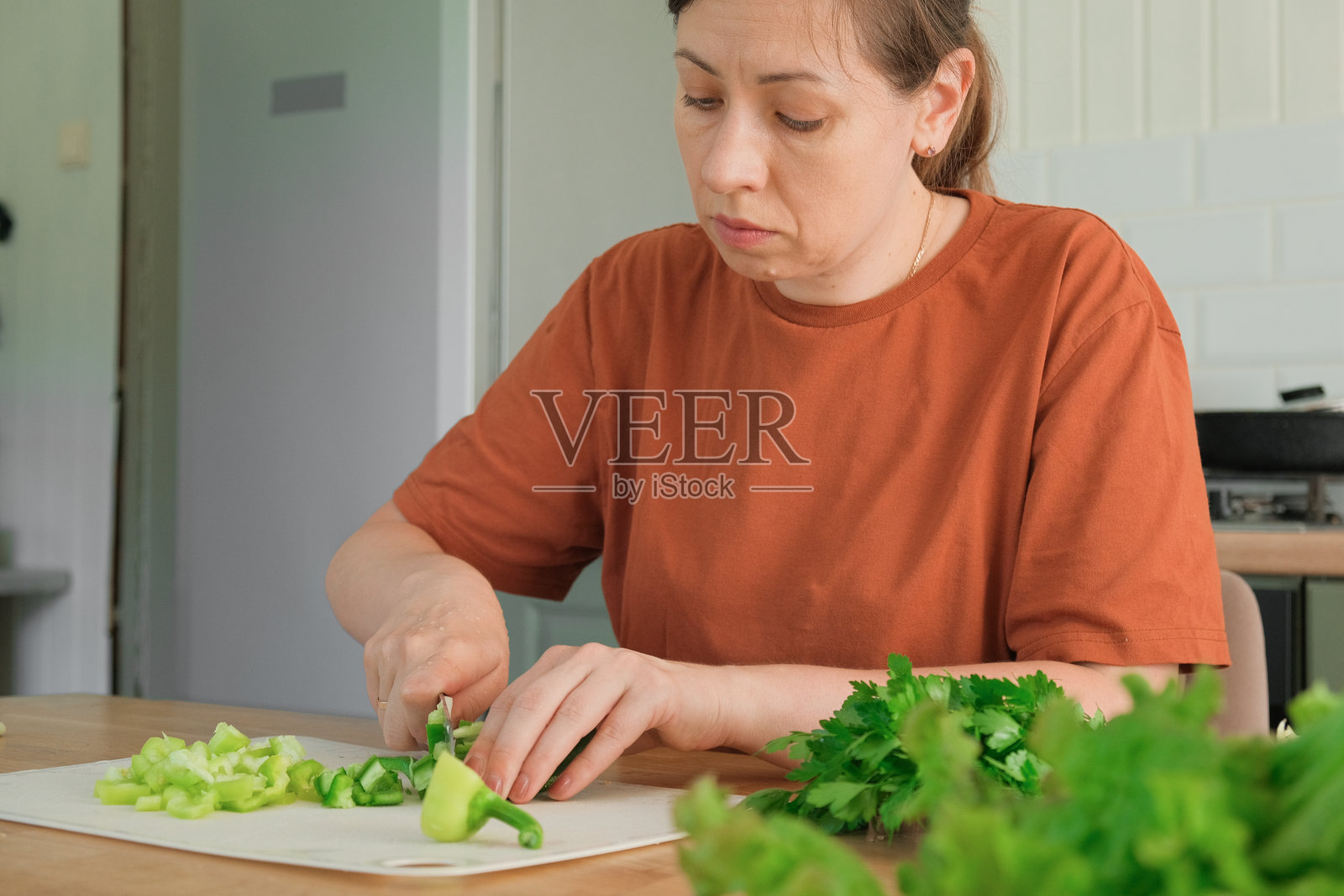 中年女性穿着赤陶色T恤，在家里的厨房里准备新鲜的绿色纯素沙拉。她正在切菜板上用刀切蔬菜和生菜，烹饪餐食。健康的饮食食物。照片摄影图片