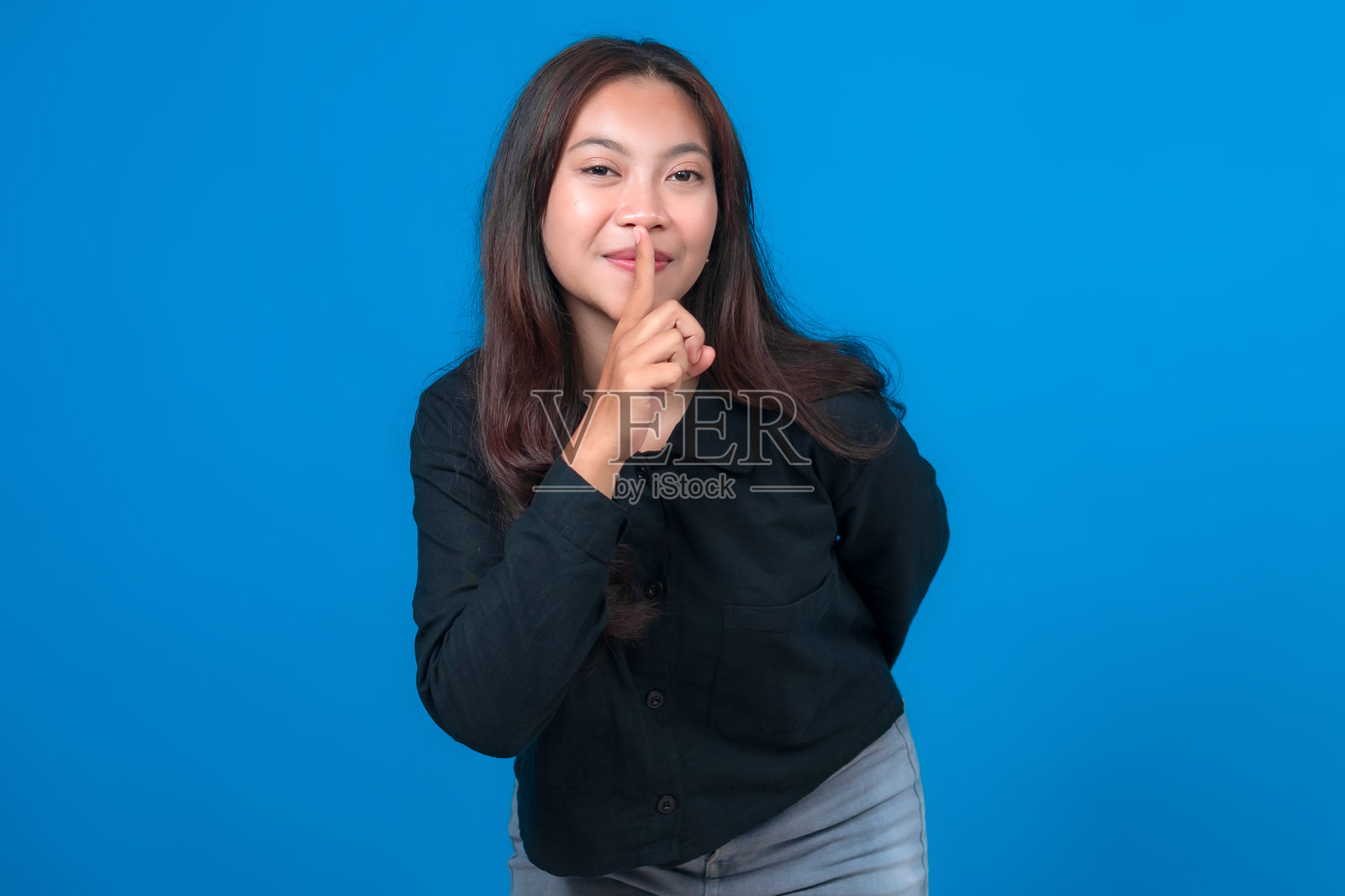 一位留着深色长发的年轻亚洲女性，身穿黑色上衣和灰色裤子，站在蓝色背景前，面带温柔的微笑，并做出嘘声手势，示意保持安静或保密。照片摄影图片