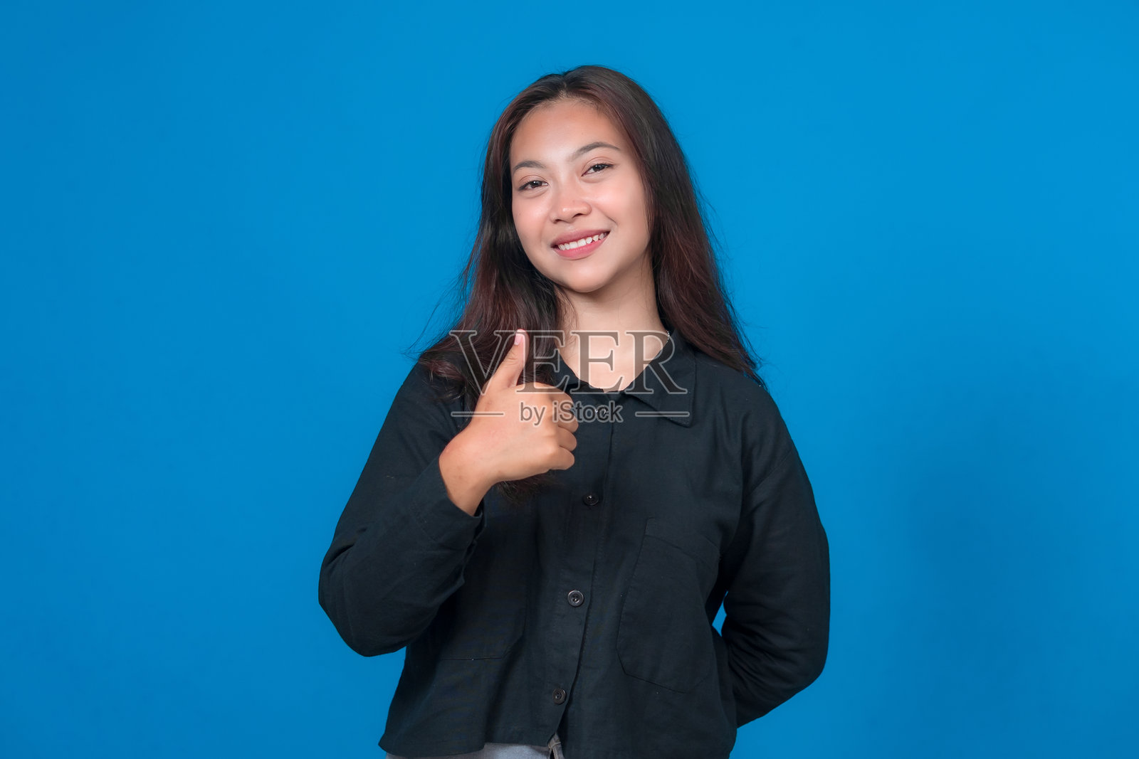 一位自信的年轻亚洲女性，带着友好的微笑，竖起大拇指，站在蓝色背景前。这个积极的手势表达了赞许、同意或鼓励。照片摄影图片