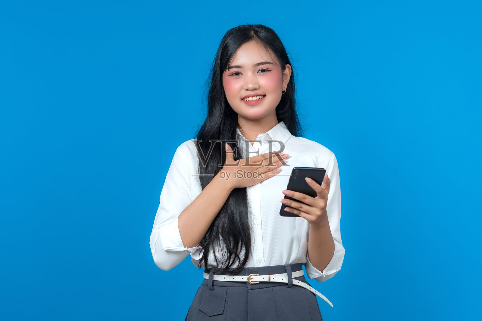 一位年轻的亚洲女性肖像，她微笑着，一只手放在胸前，另一只手拿着智能手机，表达感激或赞赏。她自信地站在蓝色背景前。照片摄影图片