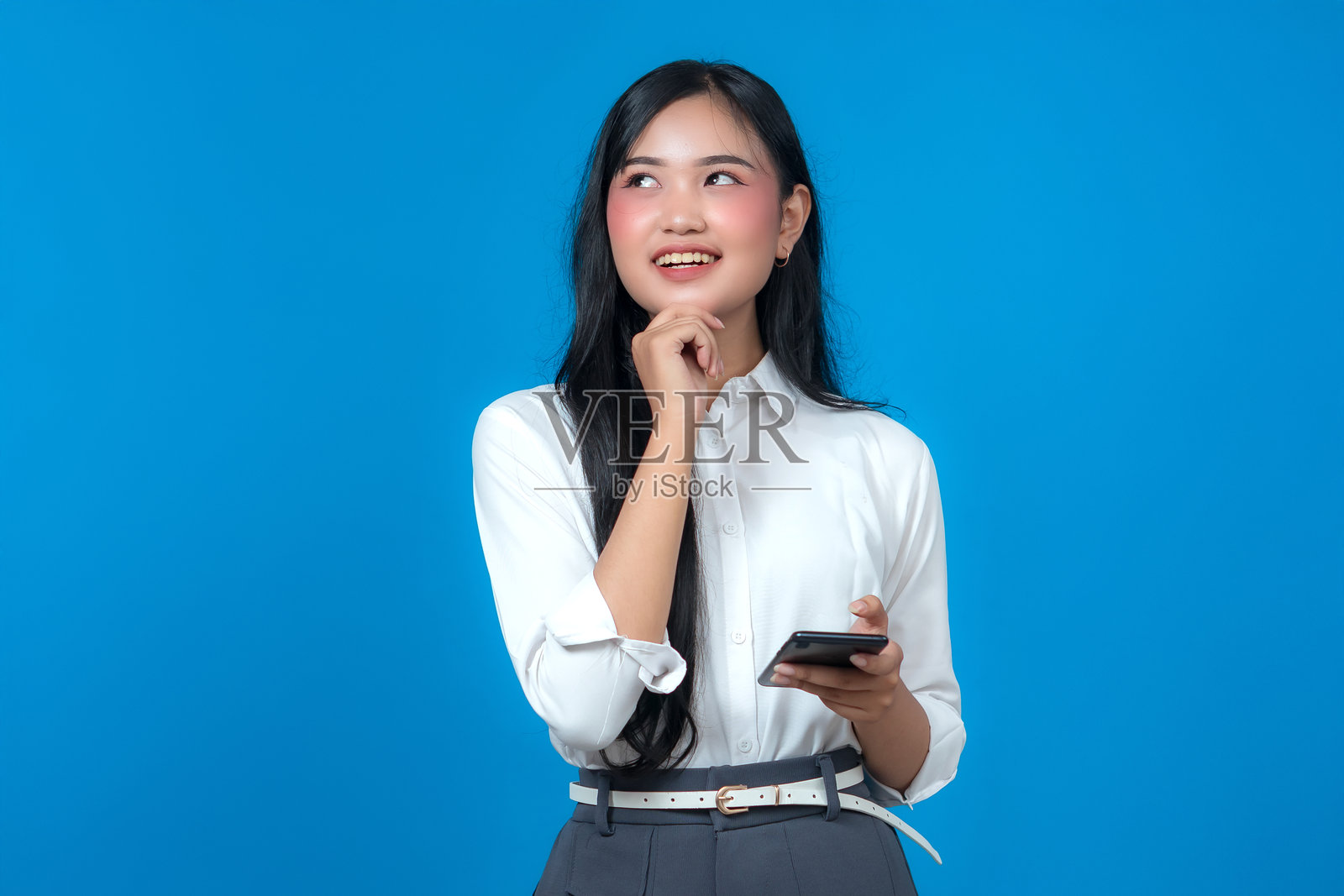 一位穿着白色衬衫的年轻亚洲女性，手持智能手机，面带微笑，带着思考的表情向上看，自信而好奇地站在纯蓝色背景前。照片摄影图片