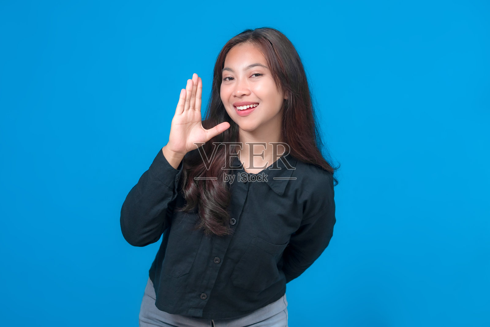 一位快乐的亚洲女性站在明亮的蓝色背景前，自信地微笑，并举起手致以友好的问候，象征着积极和亲和力。照片摄影图片