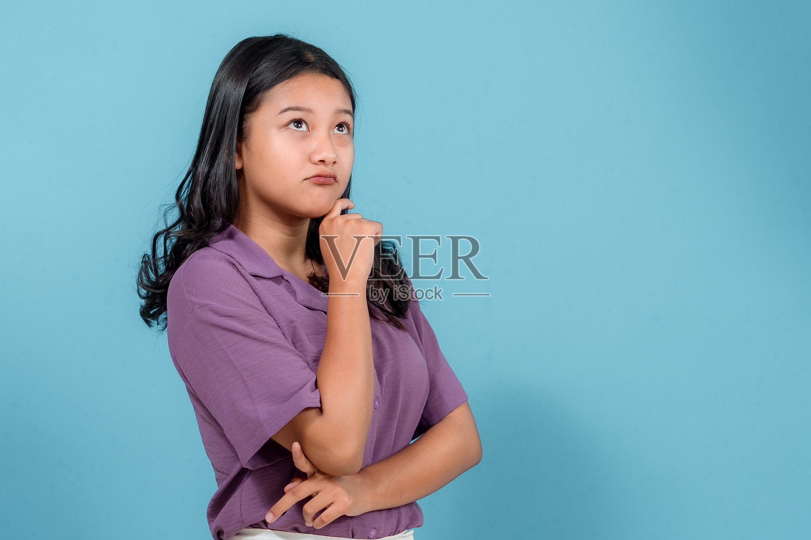 一位留着长黑发的年轻亚洲女性，在蓝色背景前若有所思地摆姿势，她抬头触摸着下巴，表达出好奇和深深的沉思。照片摄影图片
