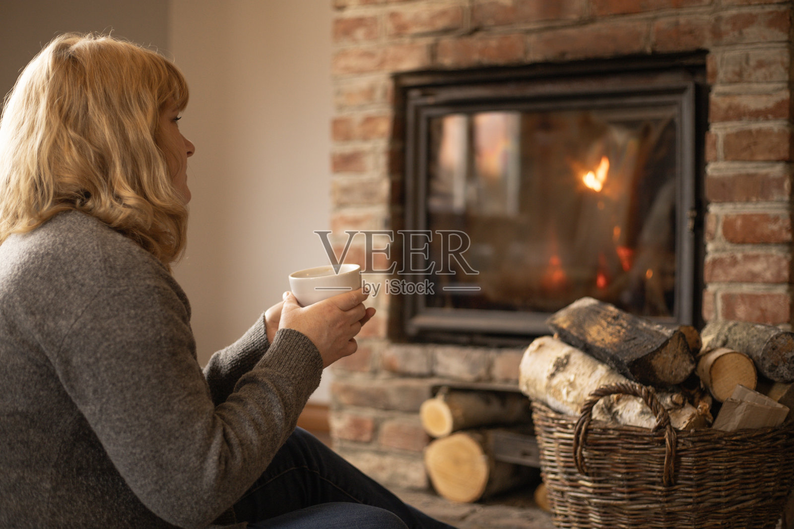 一位女士在宁静的氛围中，手持热饮，在壁炉旁静静地看着火焰。照片摄影图片