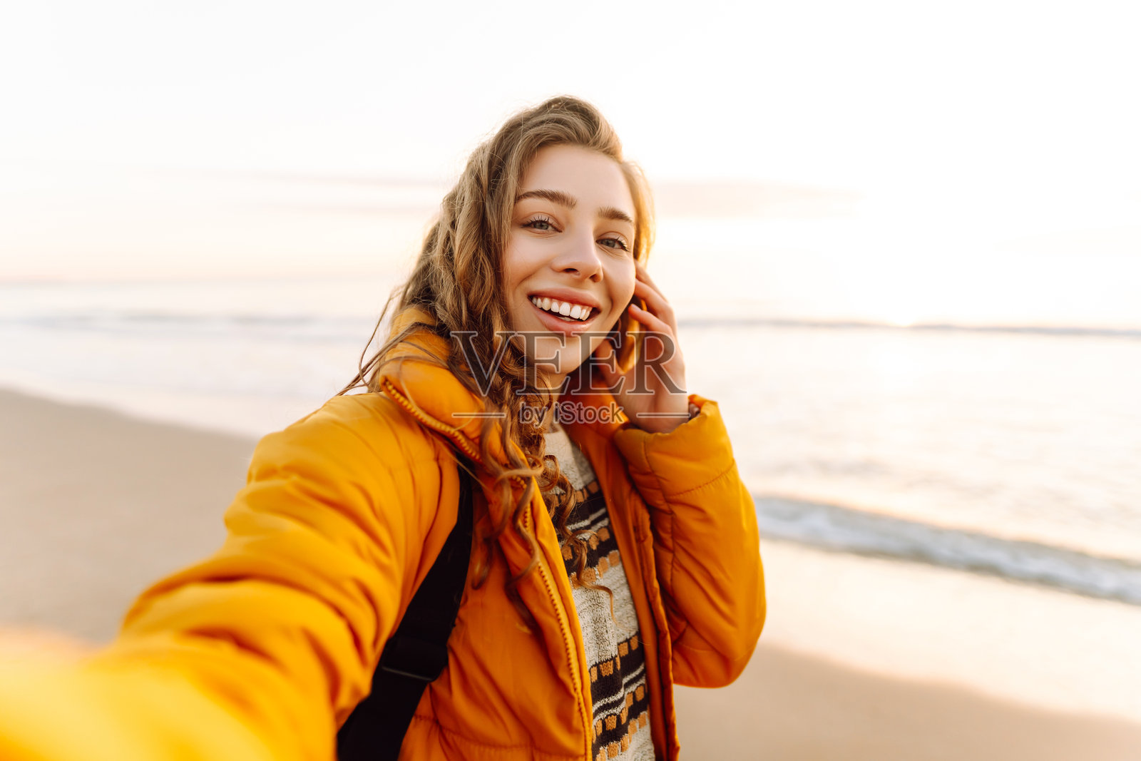 一位开朗的女士背着背包，手持手机，在日落时分的沙滩上自拍。照片摄影图片