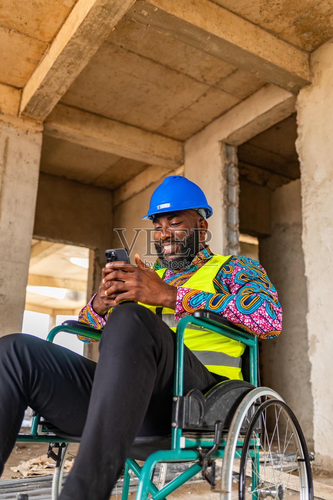 非洲建筑工程师戴着安全帽和安全背心，坐在轮椅上使用智能手机进行视频通话、购物、数字银行、社交媒体和工地上的互联网浏览。照片摄影图片