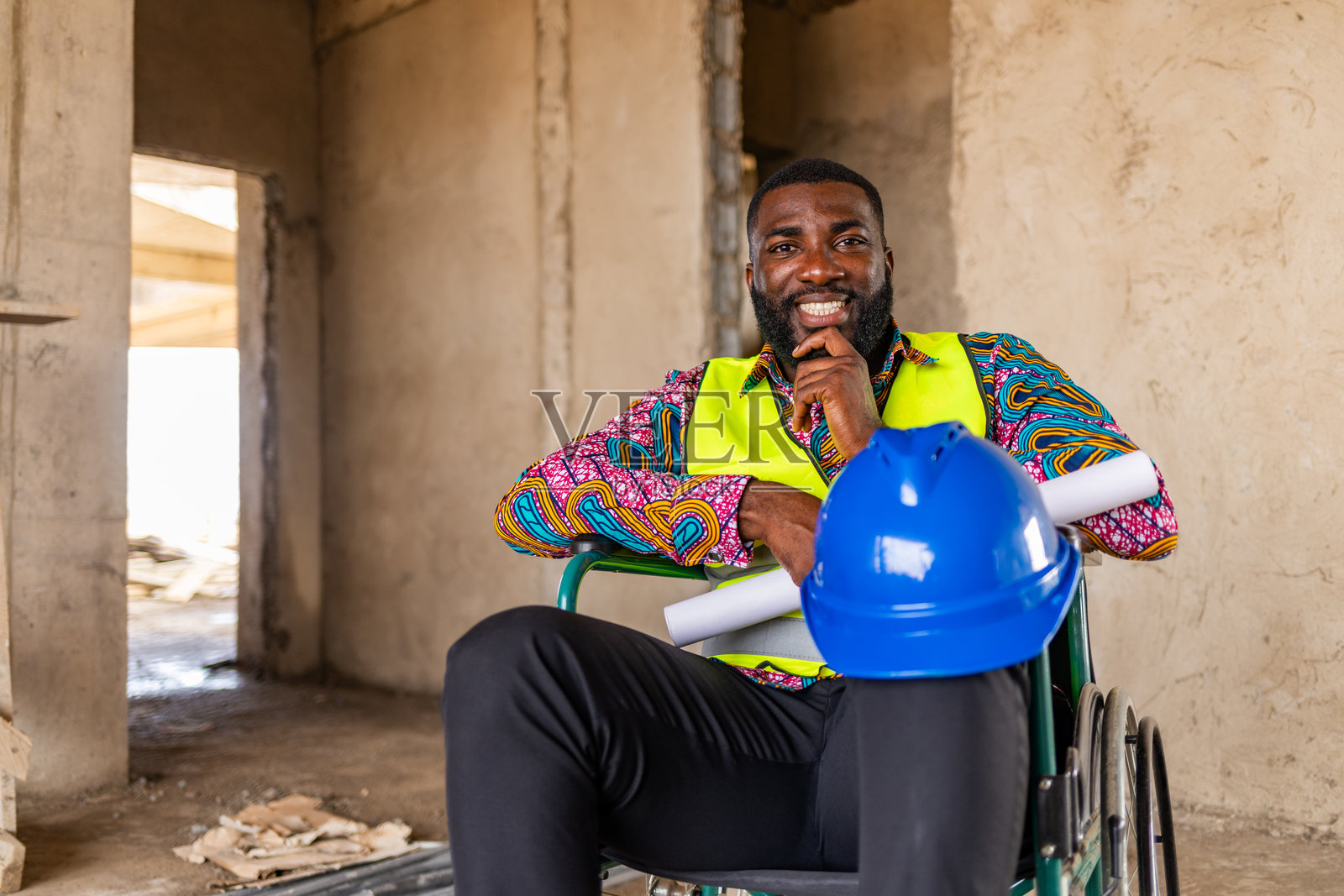 坐轮椅的非洲裔建筑工程师面带微笑，手持建筑蓝图和安全帽，在建筑工地上展现出自信。照片摄影图片