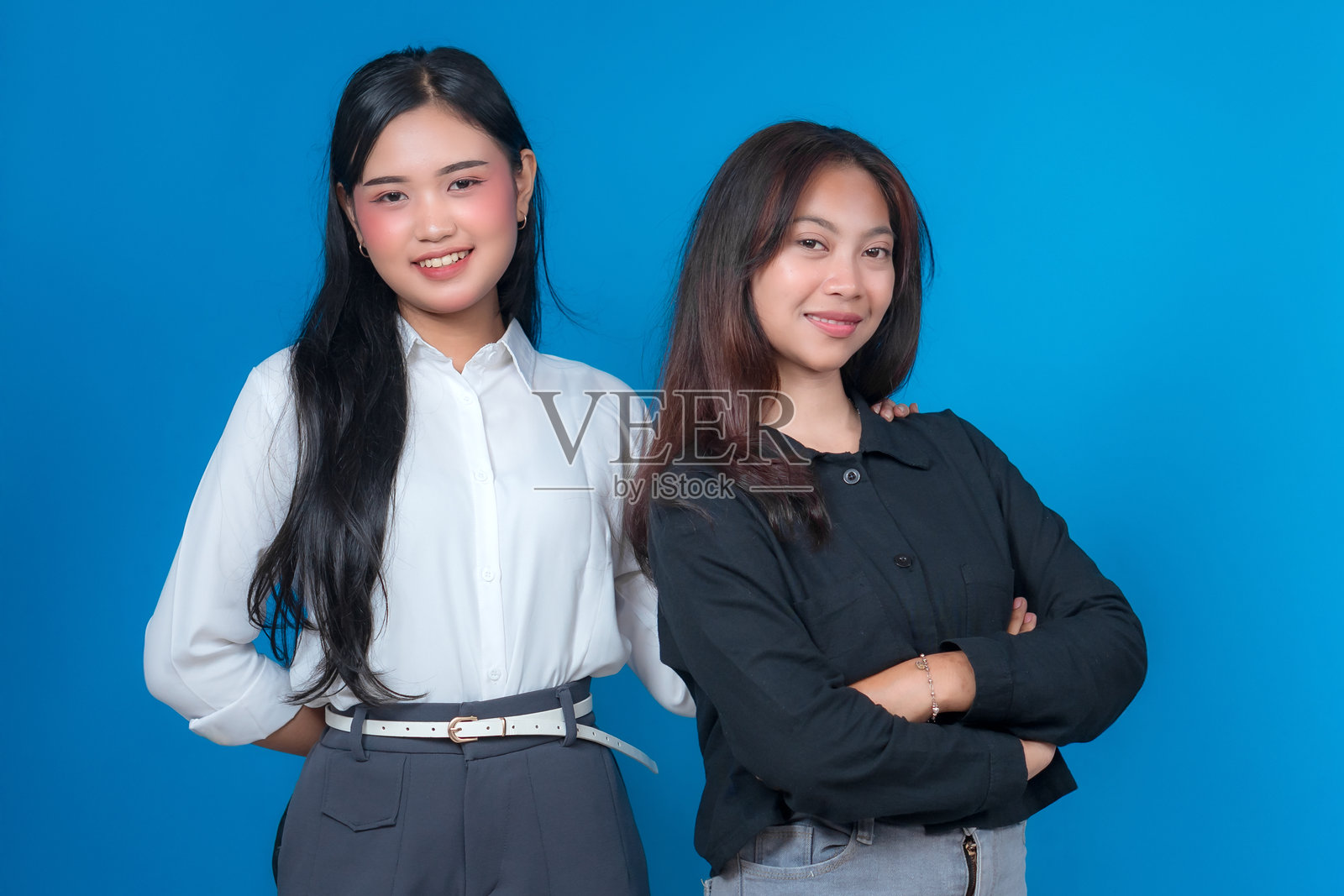 两位年轻的亚洲女性并肩站立，脸上洋溢着温暖的笑容。其中一位自信地交叉着双臂，另一位则温柔地将手搭在她的肩上，象征着支持与团结。照片摄影图片