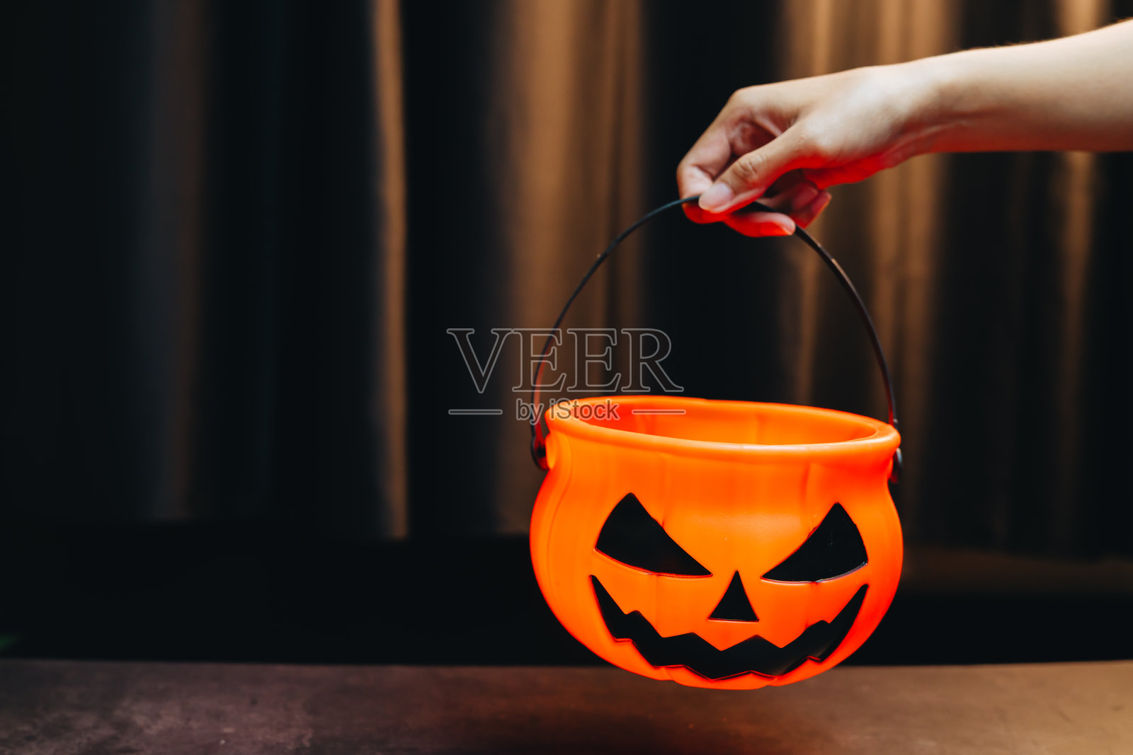 万圣节概念。一只手拿着一个带有黑色南瓜灯笑脸的橙色万圣节桶，背景为深色，用于节日装饰照片摄影图片