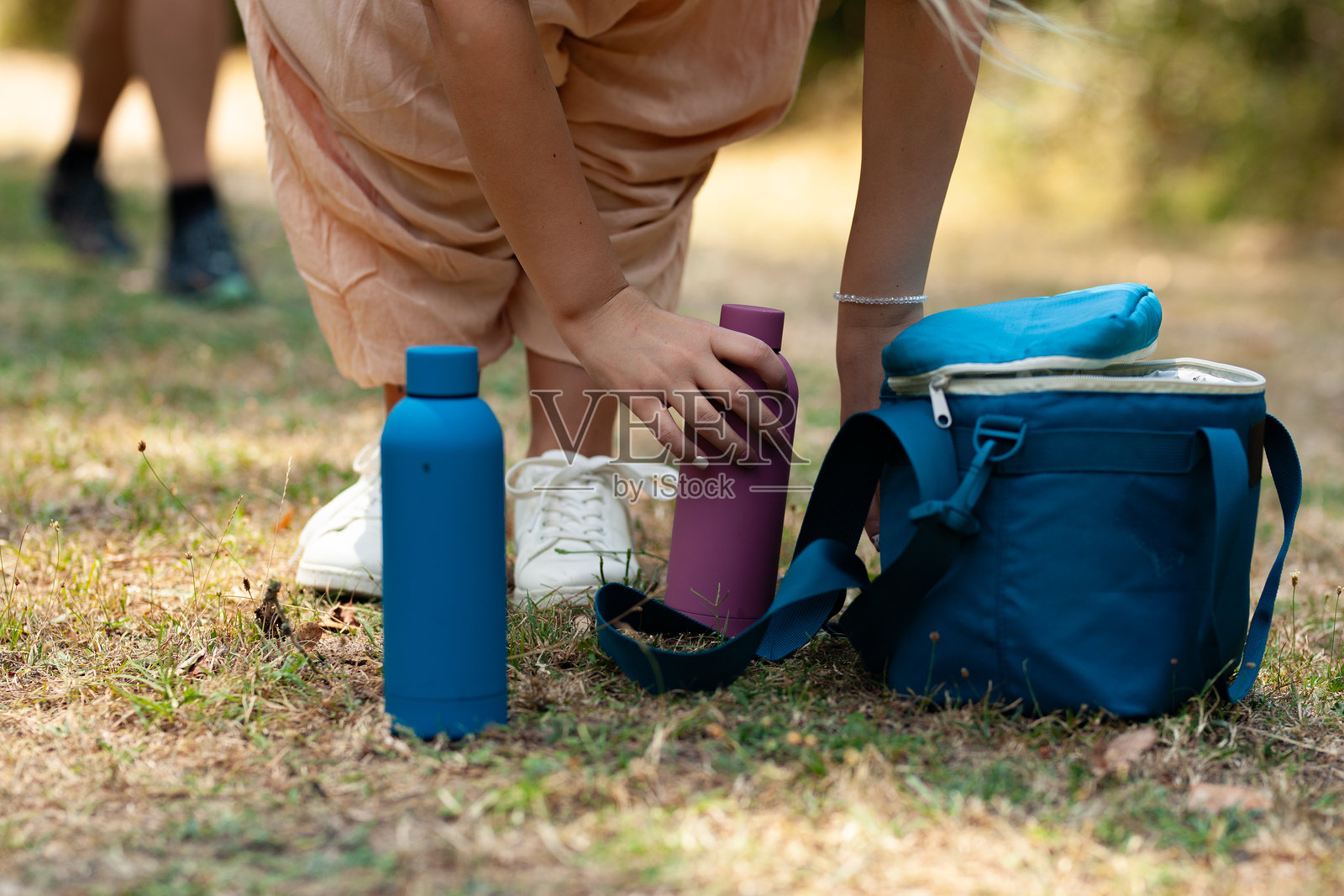 一个女孩跪在草地上，整理她五颜六色的水瓶和小包，与朋友们一起露营。背景是柔和的绿意和轻松的氛围。照片摄影图片