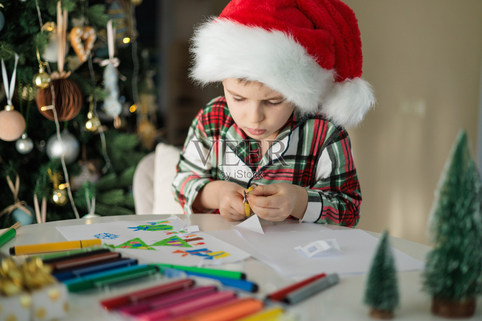 小男孩戴着圣诞帽，在家里的圣诞树旁，用纸和马克笔制作节日手工艺品照片摄影图片
