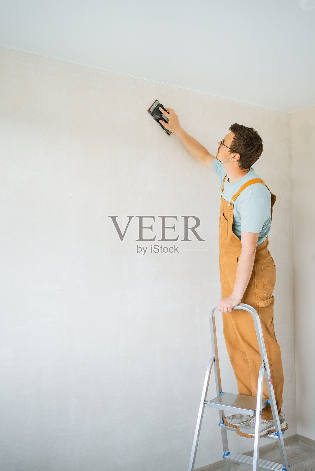 穿着米色工装的建筑工人在金属梯子上保持平衡，用手动砂光机打磨白墙，为光滑的油漆和翻新做准备照片摄影图片