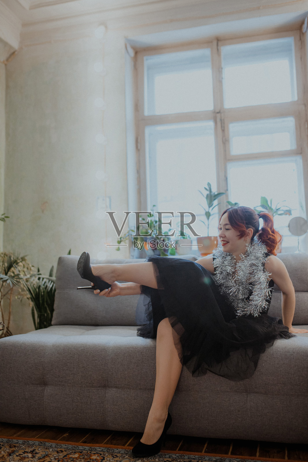 时尚女子身着黑裙，佩戴闪光饰物，在明亮的客厅沙发上整理鞋子。照片摄影图片