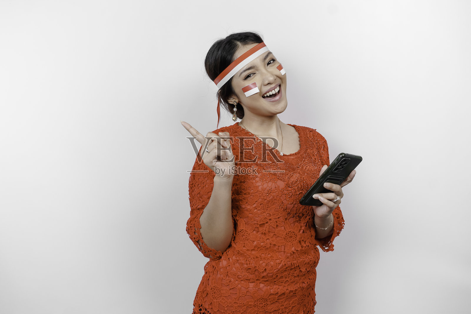 一位戴着头带、身穿红色马来装的快乐亚洲女性，拿着手机，手指指向顶部的空白区域。照片摄影图片