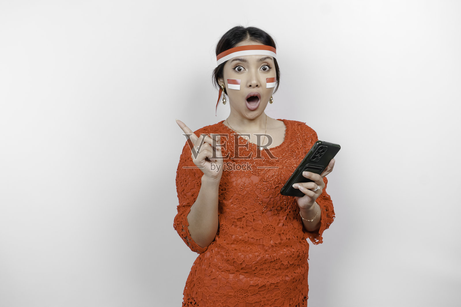 身穿红色马来装和头带的亚洲女性，表情震惊，手持手机，并指向顶部的复制空间。照片摄影图片