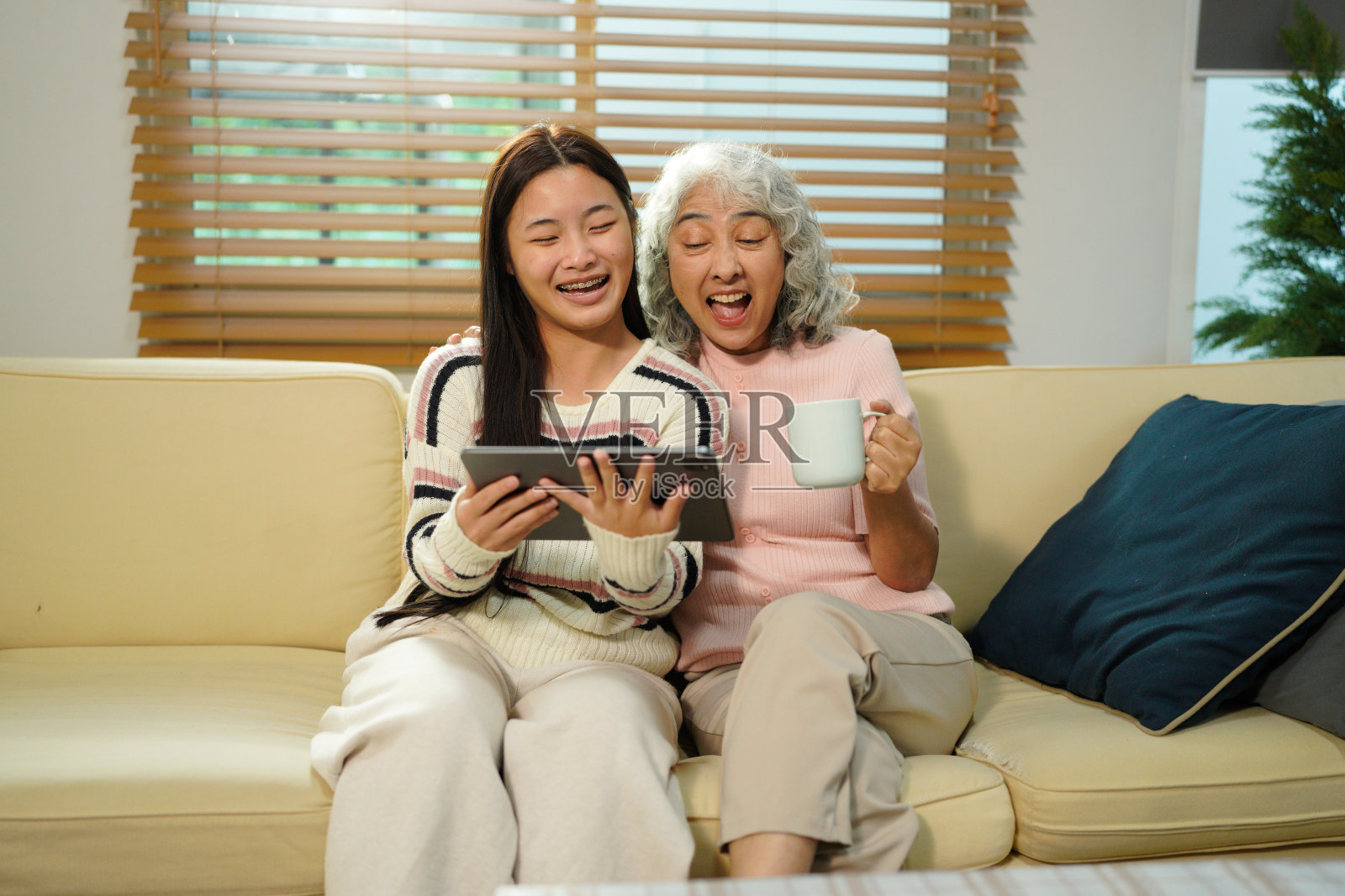 祖孙俩在家中沙发上坐着，奶奶和孙女一起使用平板电脑，脸上洋溢着笑容。照片摄影图片