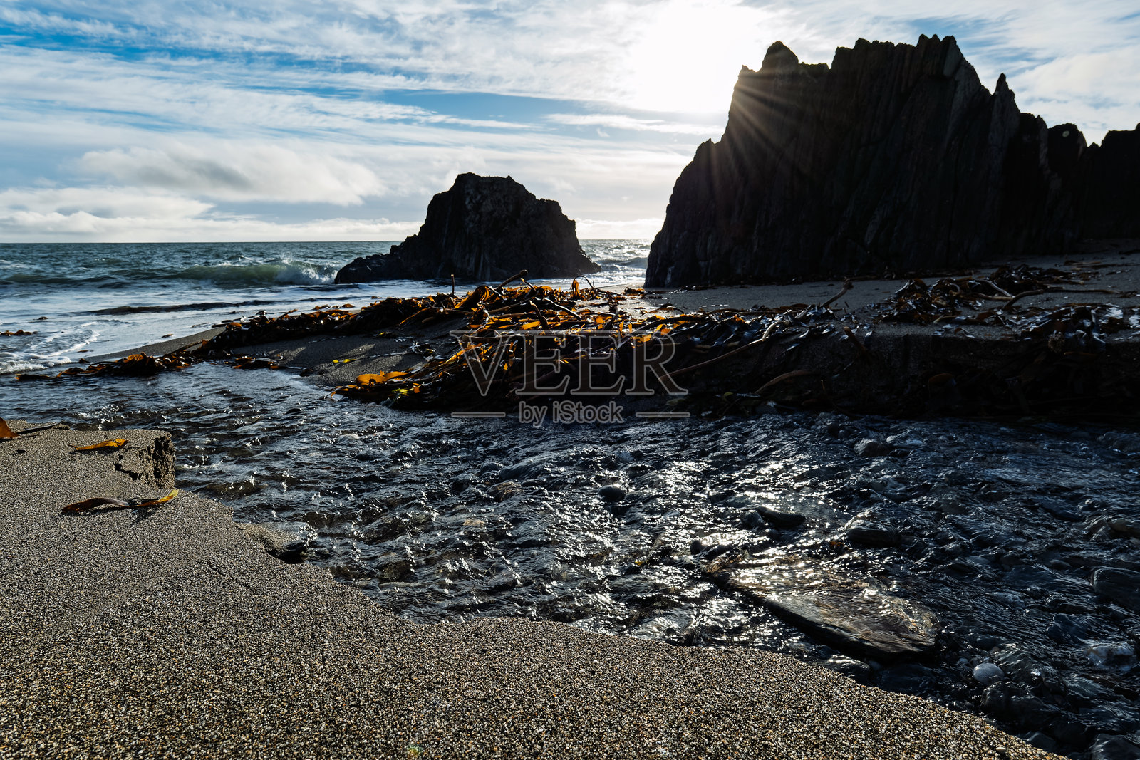 爱尔兰羊湾，一条小溪流淌到沙滩上，汇入大海。海藻散落在黑色的、锯齿状的岩石构造附近的海滩上。照片摄影图片