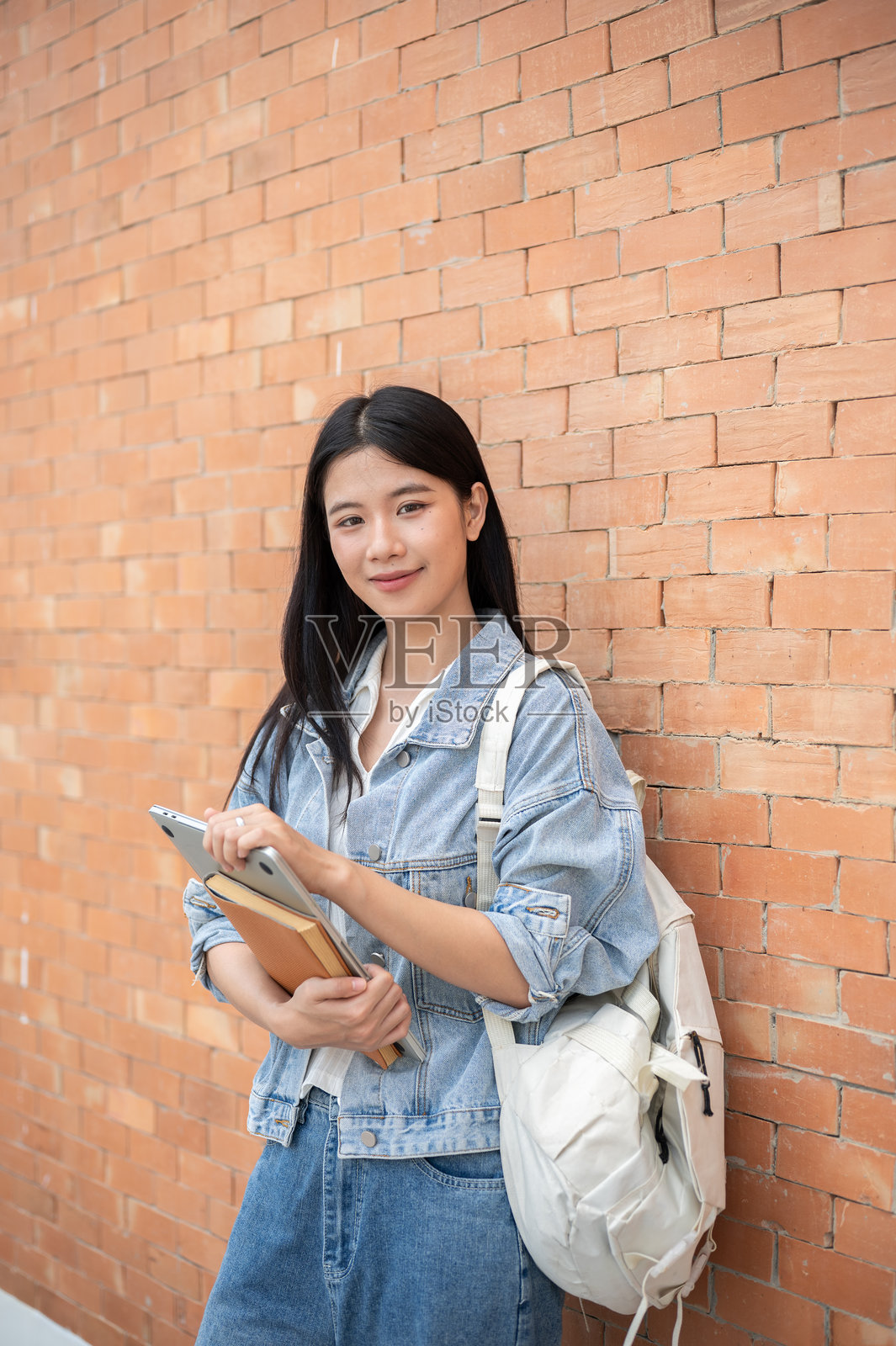 年轻的亚洲女学生背着背包，手持笔记本电脑，站立在书本上，靠着砖墙照片摄影图片