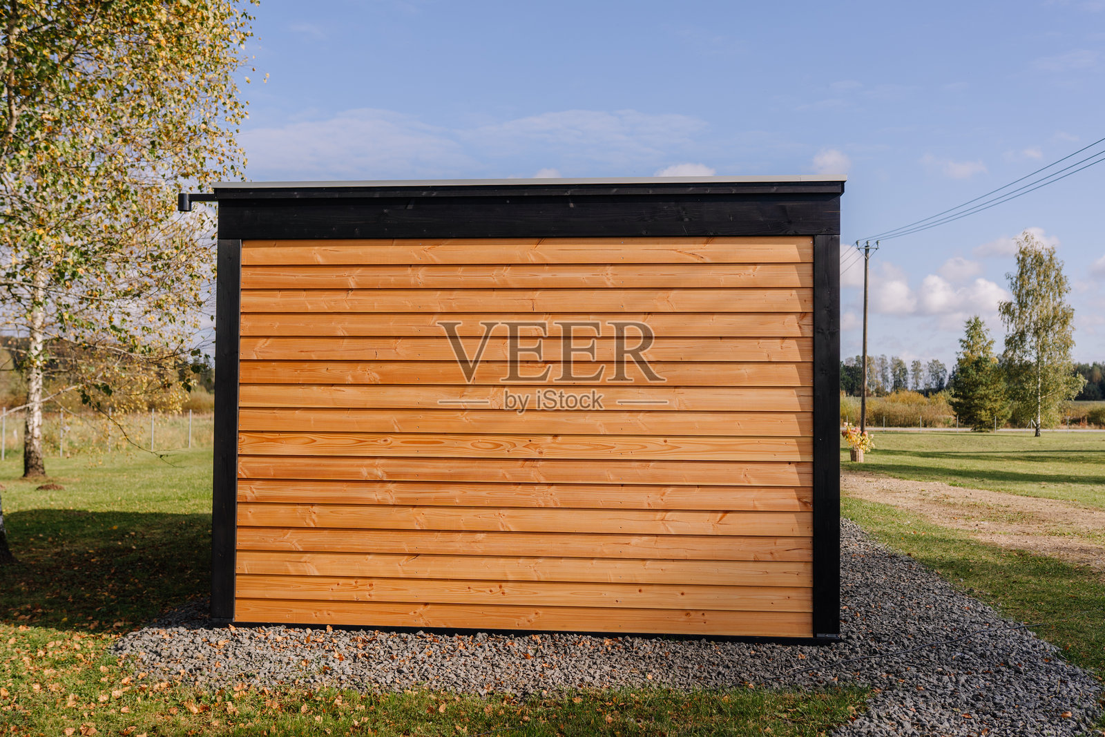 一栋现代风格的木制花园小屋侧视图，有水平壁板、黑色饰边和砾石地基，坐落在一片蓝天下葱郁的乡村景观中。照片摄影图片