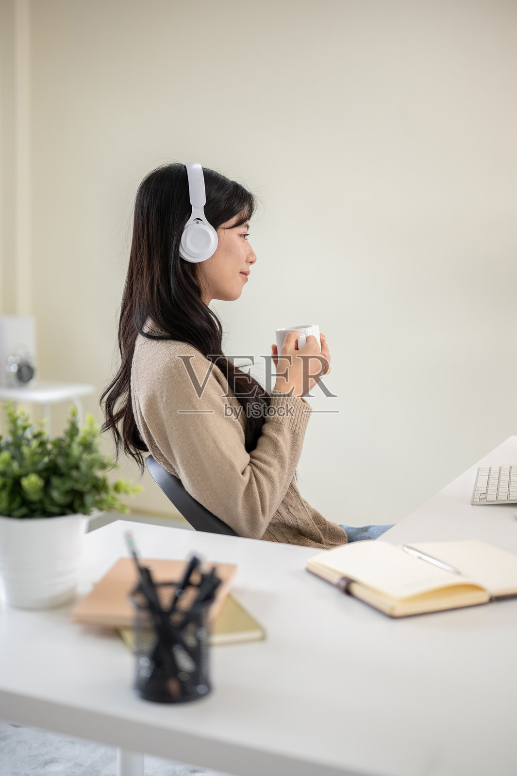 一位戴着耳机、手持咖啡杯的亚洲女性，她正坐在办公室的桌子旁或客厅里。照片摄影图片