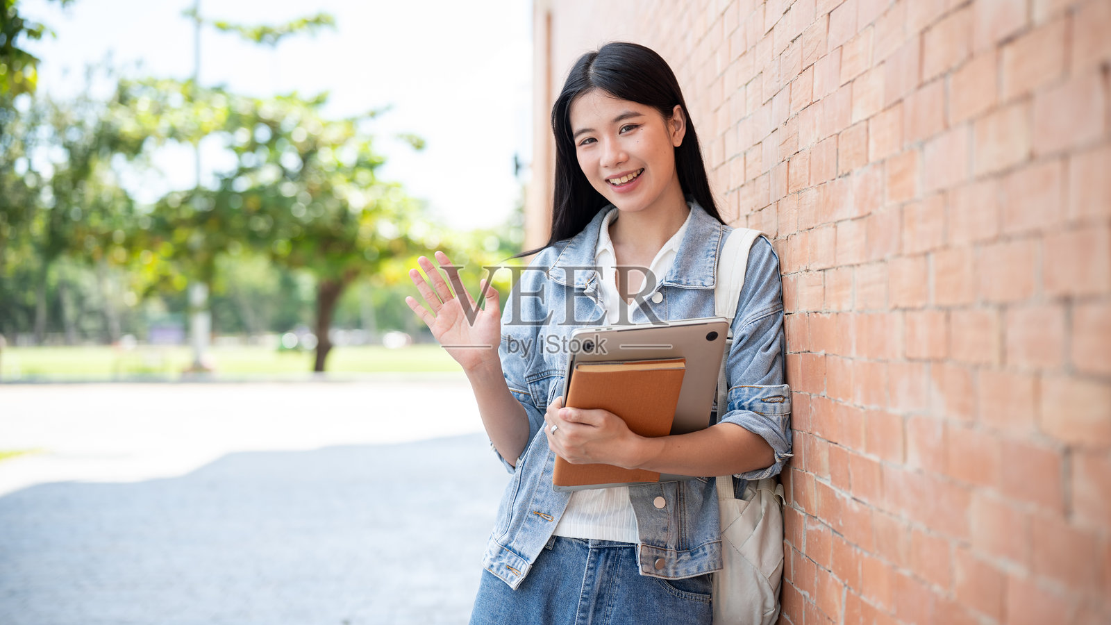 年轻的亚洲女学生，手持笔记本电脑和书籍，靠在砖墙上挥手。照片摄影图片