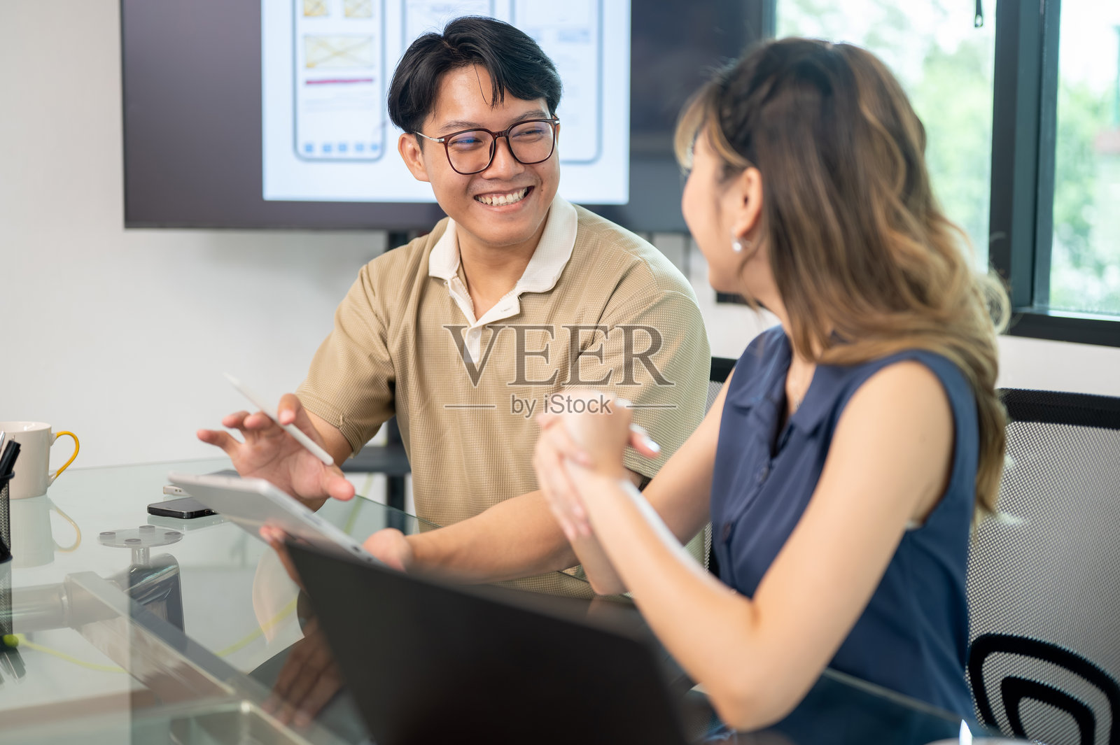 戴眼镜的男人微笑着，手里拿着平板电脑和触控笔，在办公室的会议室里与女同事交谈。照片摄影图片