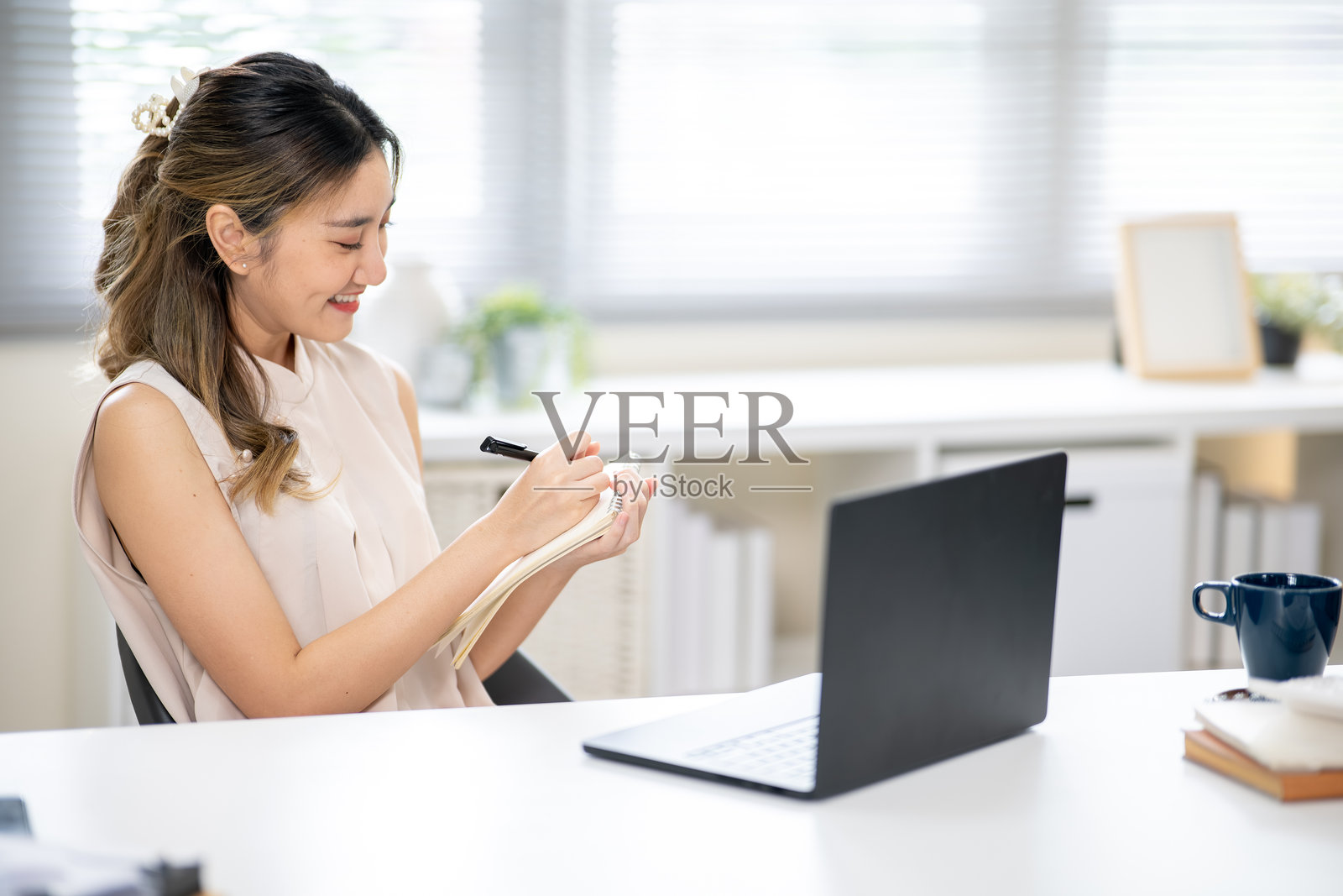 办公室里，一位职业女性在白色桌子上，笔记本电脑旁，手持笔记本并用笔书写。照片摄影图片