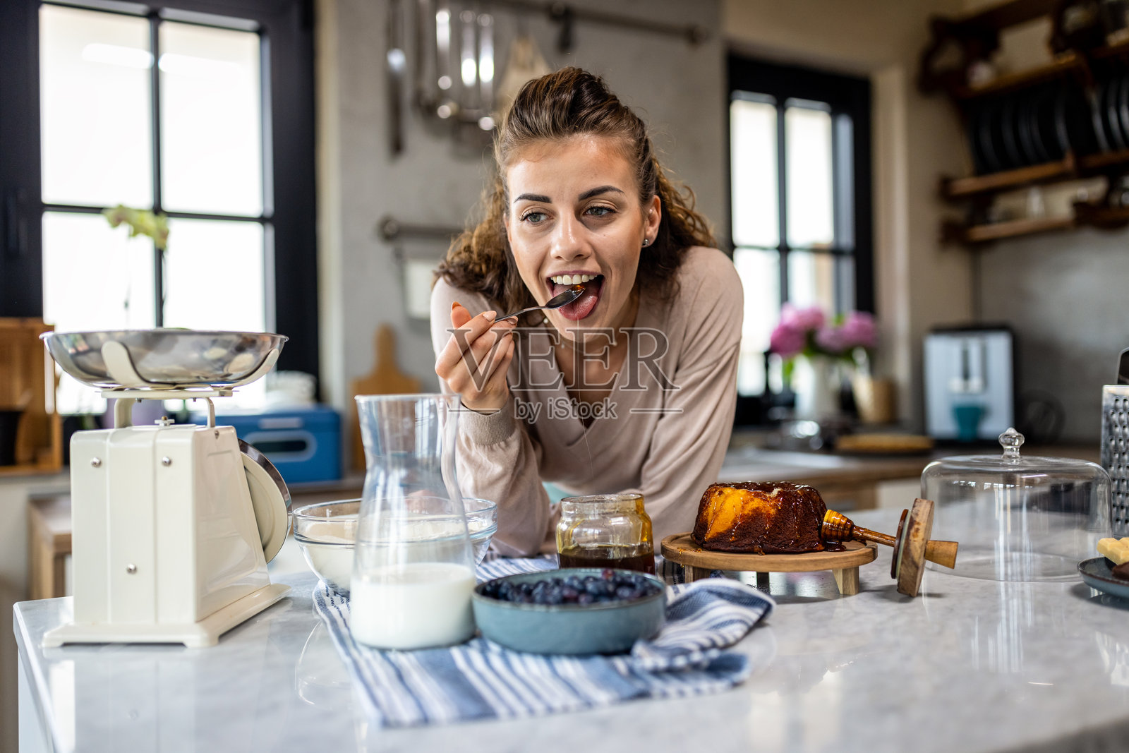 女人在明亮的厨房里品尝蛋糕，旁边有牛奶、一罐果酱和新鲜的糕点照片摄影图片