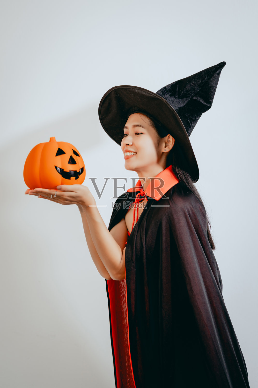 一位穿着女巫服装的美丽亚洲年轻女性，手持橙色南瓜，在干净的白色背景前欢庆万圣节，散发出节日气氛、快乐和俏皮的假日魅力。照片摄影图片