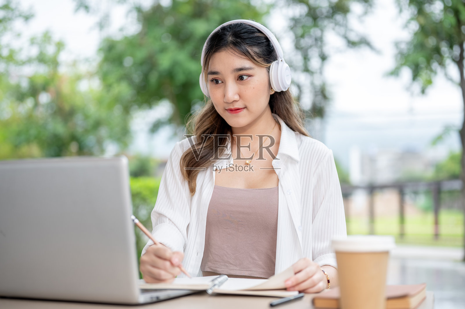 年轻的亚洲女性戴着耳机，手持铅笔在笔记本上书写，同时看着公园桌子上的笔记本电脑。照片摄影图片