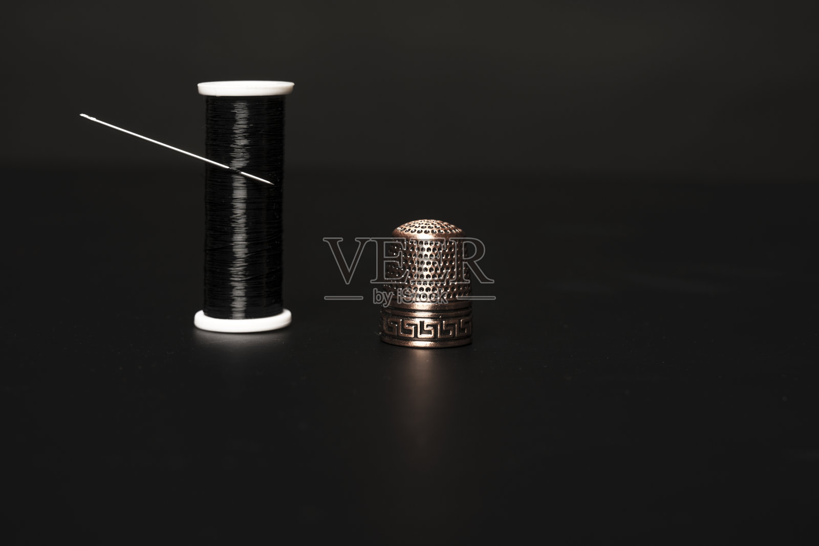 黑色背景上，一个孤零零的闪亮黑色线轴旁边放着一根缝纫针和一个铜顶针照片摄影图片