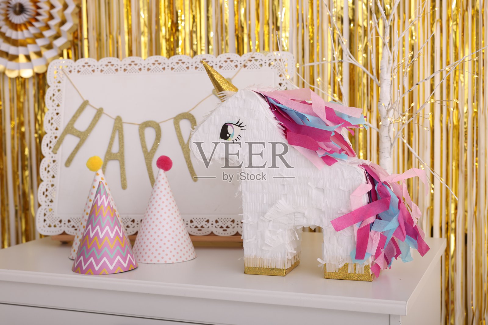 室内白色抽屉柜上摆放着造型为独角兽的明亮派对 पिñata 和派对装饰品，背景是锡箔流苏窗帘照片摄影图片