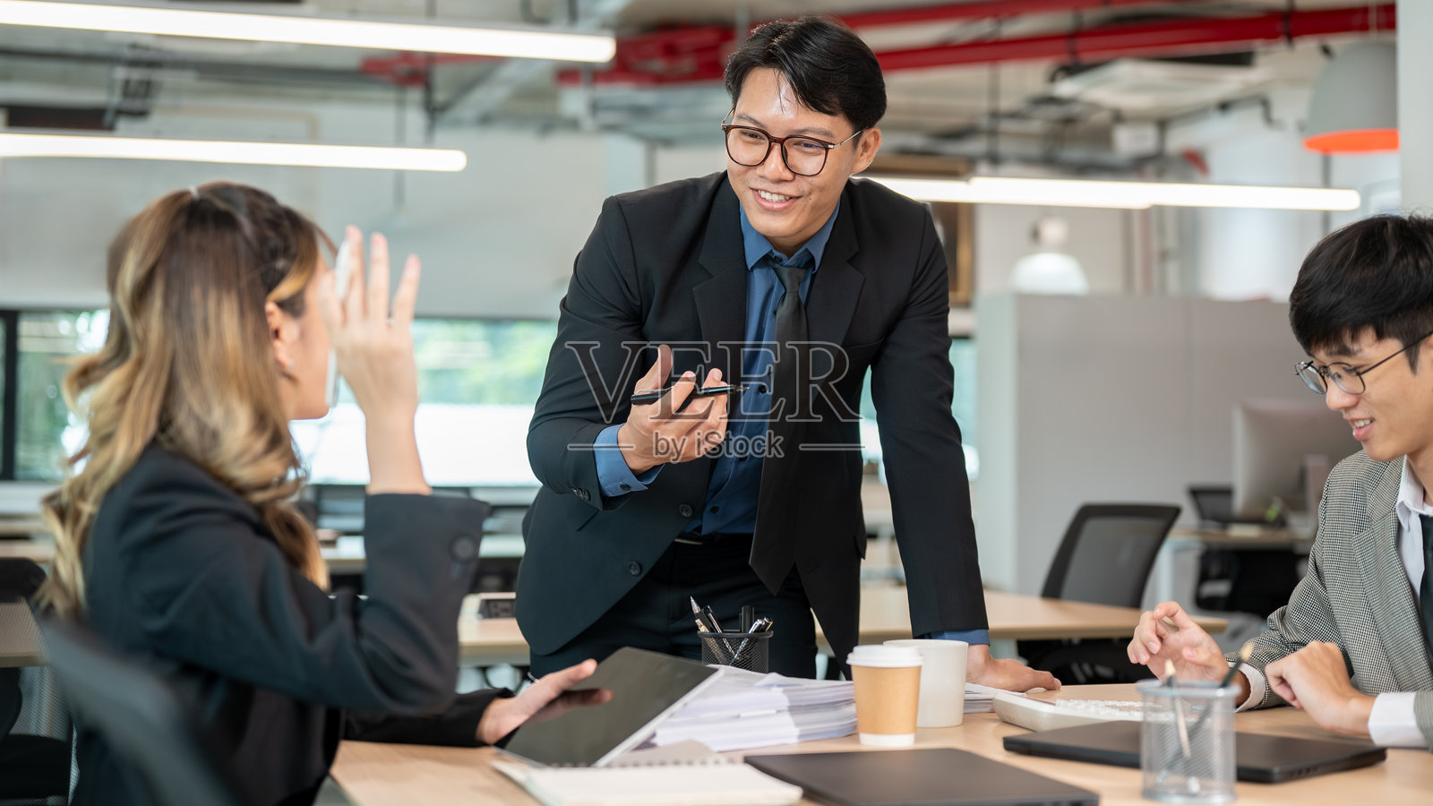 在办公室里，一位男商人拿着笔，与一位举手提问的女同事在木桌旁交谈。照片摄影图片