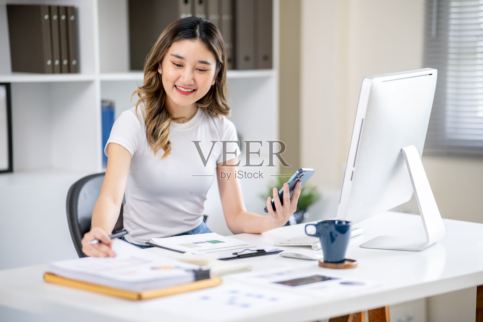 一位亚洲女办公室职员坐在电脑桌前，一手拿着手机，一手看着文件。照片摄影图片