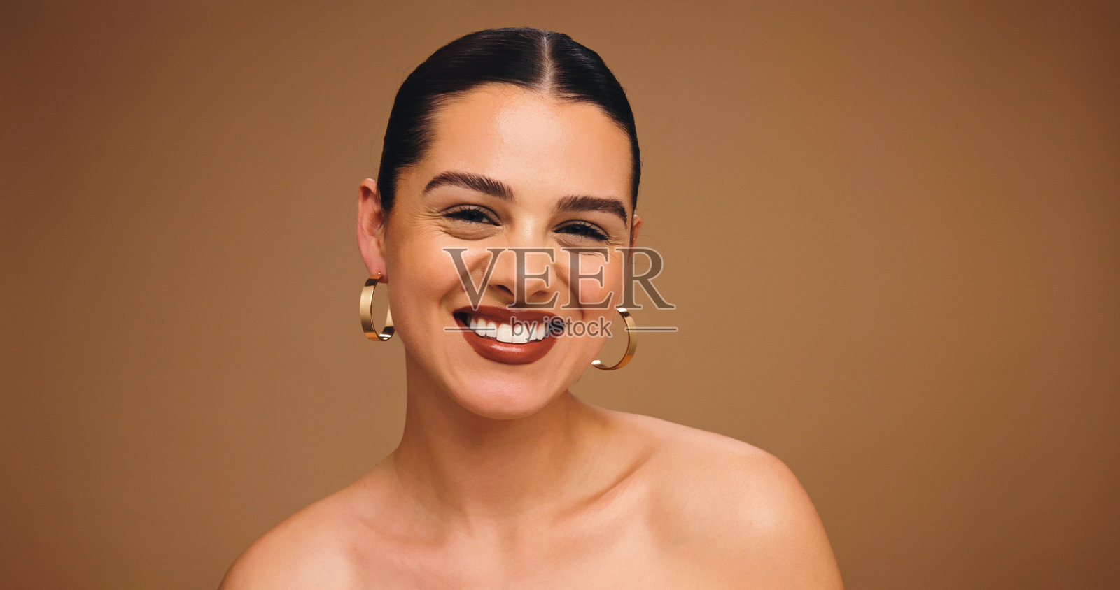 工作室里，一位快乐的女士化着妆，戴着耳环，在棕色背景的空间中展现出迷人的光彩和光泽。这是关于美丽、耳饰以及一位有着口红彩妆、容貌蜕变和形象改变的模特的肖像。照片摄影图片