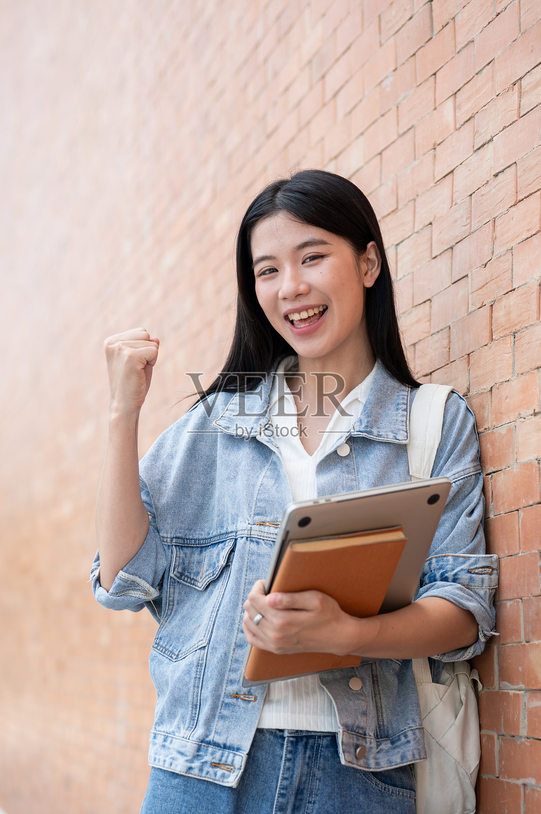 一位年轻的亚洲女学生，手持笔记本电脑和书籍，握紧拳头，靠在砖墙上。照片摄影图片