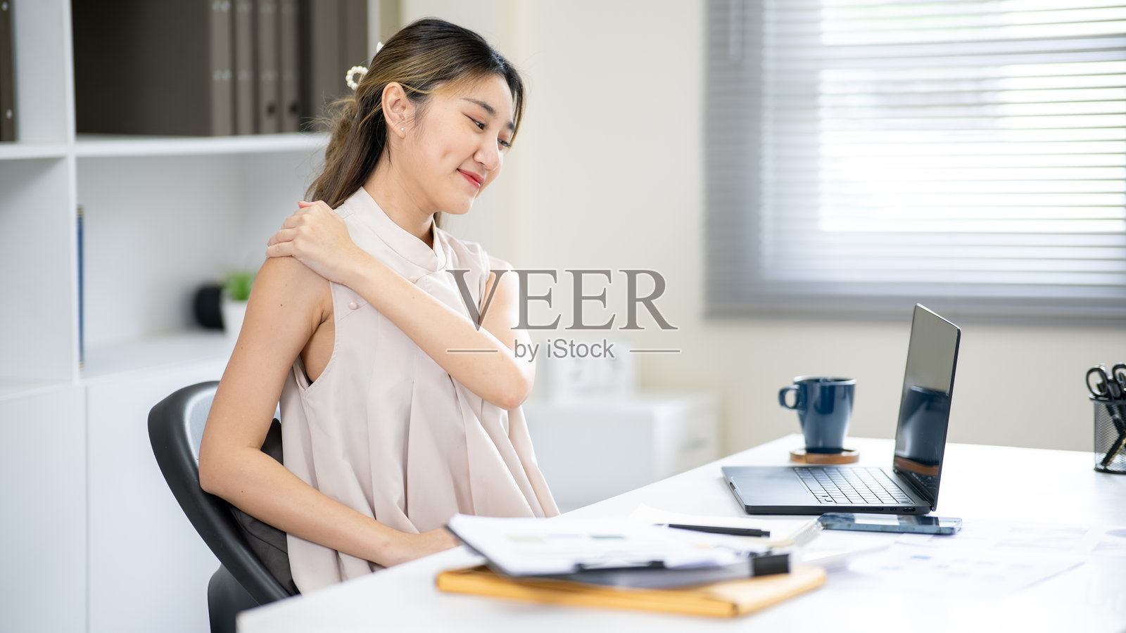 微笑的亚洲女办公室职员在办公桌前边看笔记本电脑边按摩肩膀。照片摄影图片
