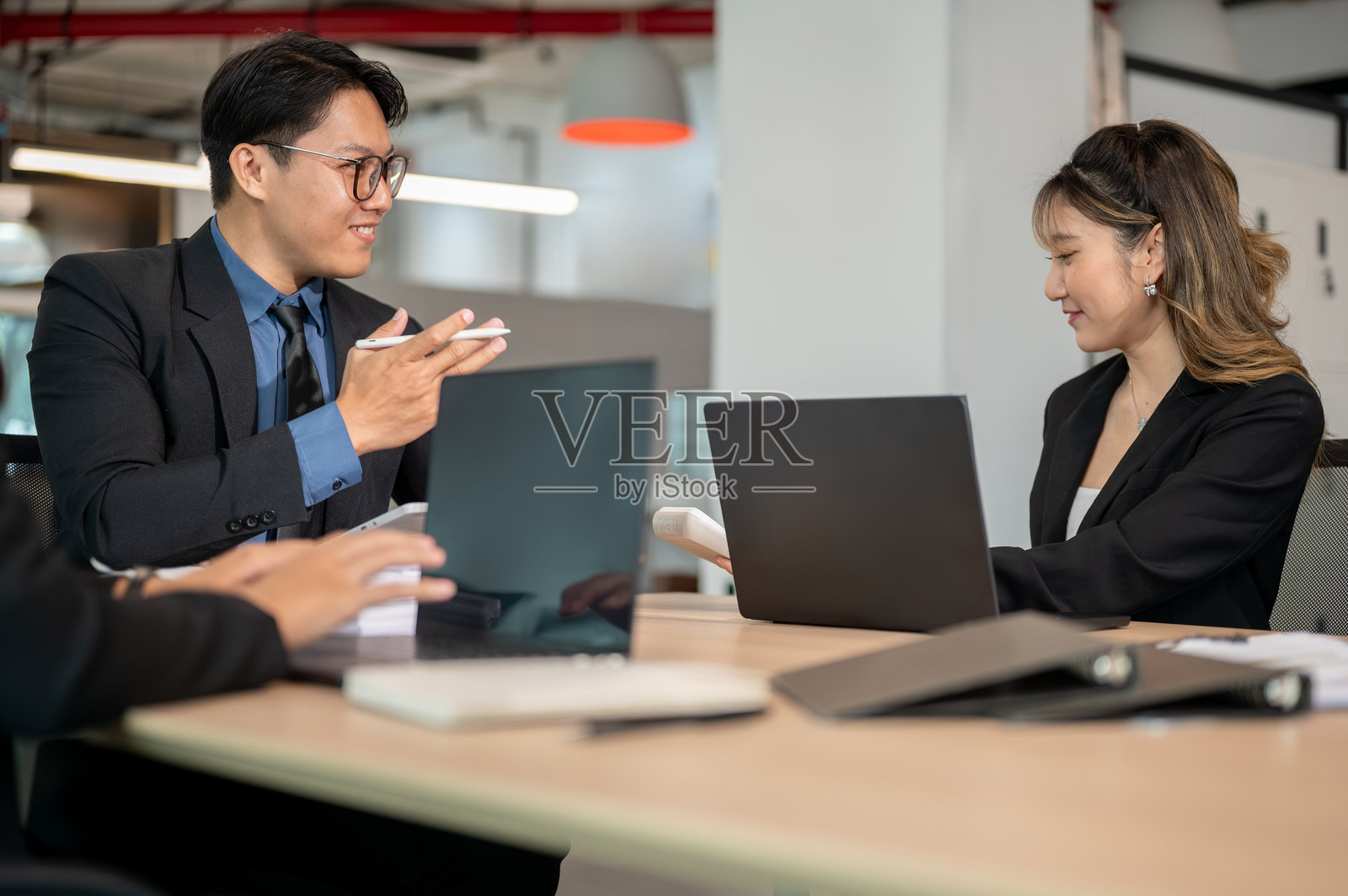 戴眼镜的商人坐在办公室的木桌旁，手持钢笔与女同事交谈。照片摄影图片