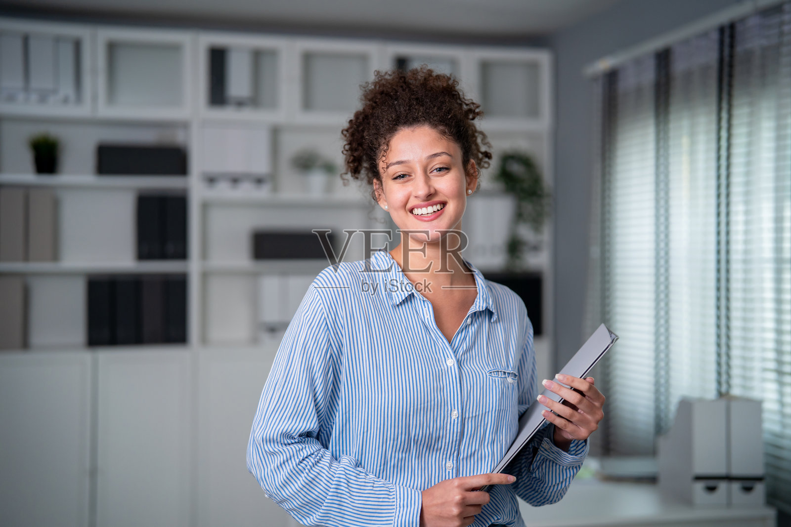 现代办公室里工作的年轻拉丁裔女商人的肖像。一位专业、漂亮的女性员工，她感到快乐放松，自信地站在办公桌旁，在舒适的工作场所看向镜头。照片摄影图片