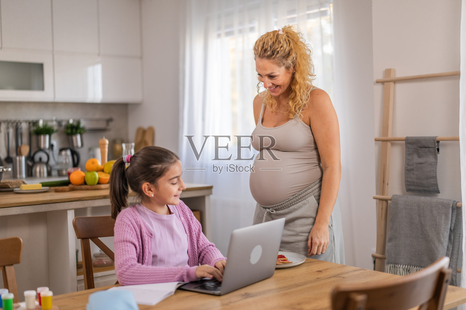 一位尽职尽责的孕妇在餐桌旁，帮助女儿在线完成家庭作业。照片摄影图片