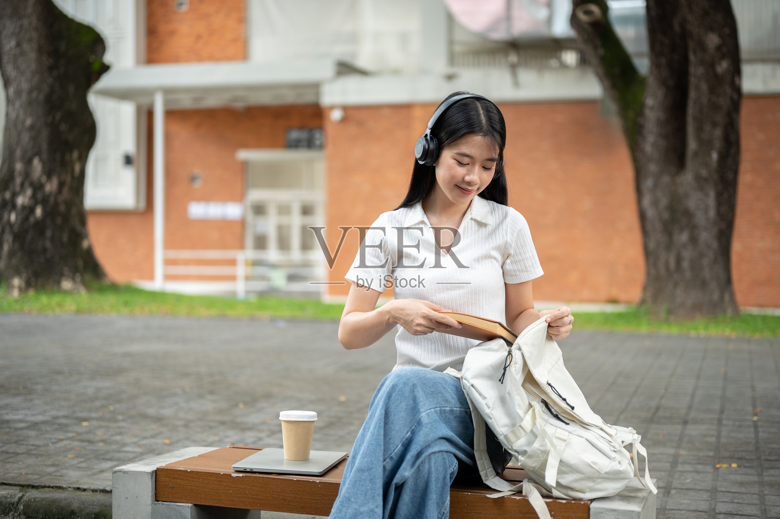年轻的亚洲女学生戴着耳机，把书放进包里，坐在人行道旁的木凳上照片摄影图片