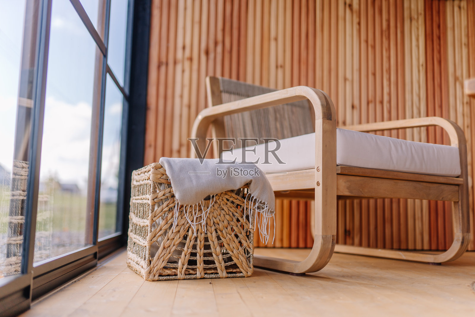 舒适的木质室内，靠近大玻璃窗，特写木扶手椅和柳条编织篮，配有浅灰色毯子，室内有自然光。照片摄影图片