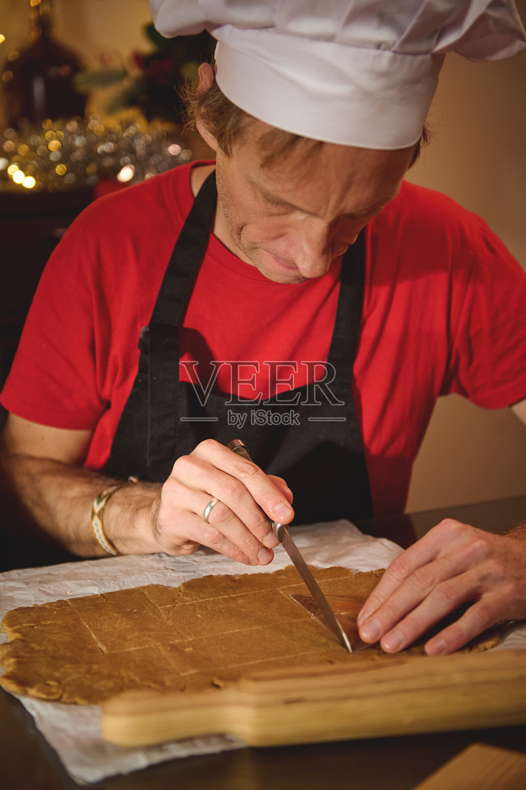 身穿红衣、系着黑围裙的厨师戴着白色厨师帽，在舒适的厨房里用烘焙纸切面团，在温馨的工作室环境中展示烘焙技巧和烹饪工艺照片摄影图片
