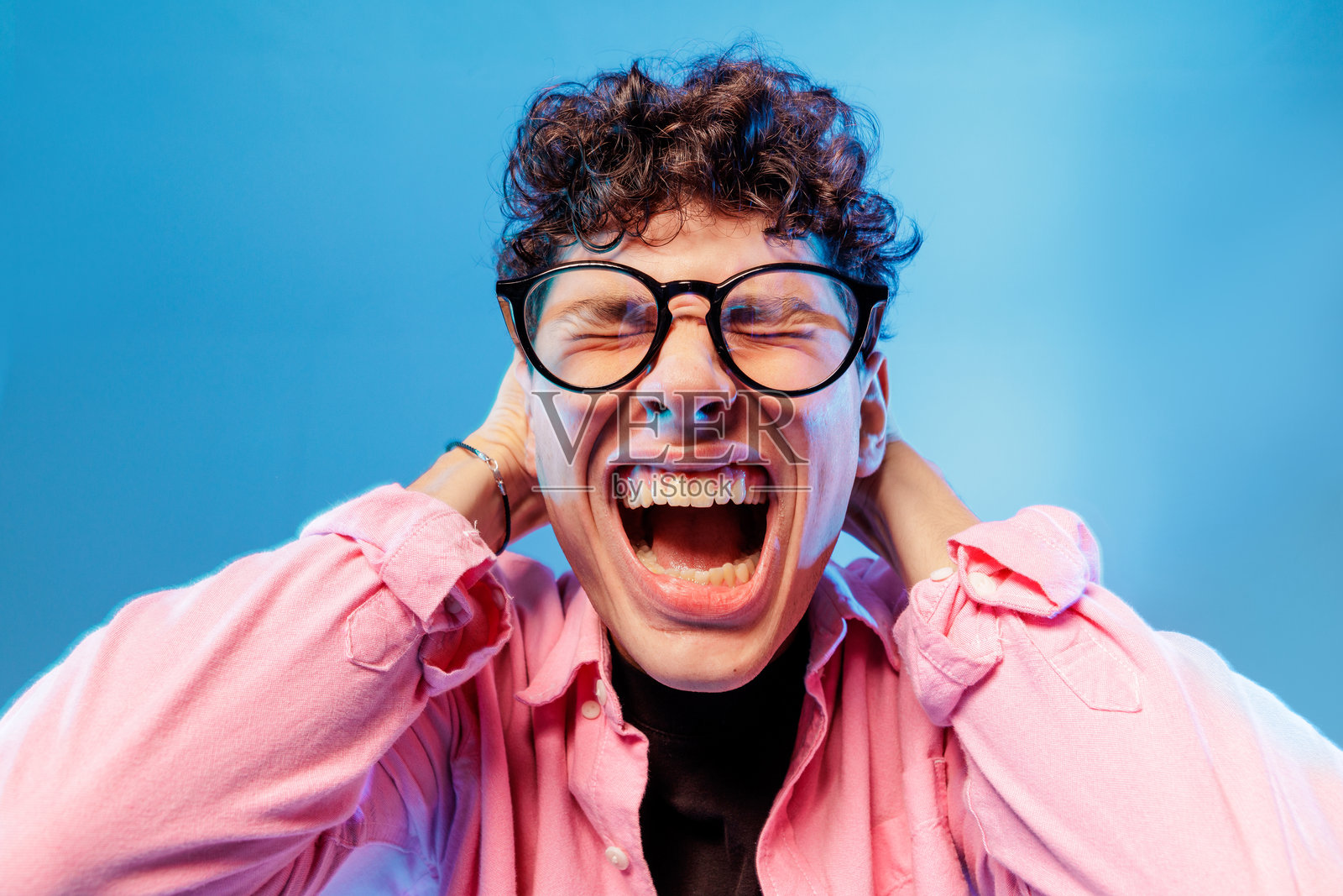 戴眼镜的青少年在蓝色工作室背景前，双手抱头尖叫，显得压力重重照片摄影图片