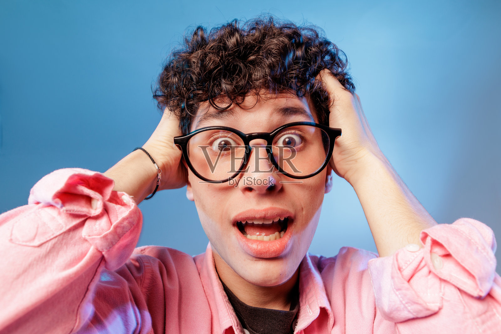 戴眼镜的震惊的青少年，双手抱头，在蓝色的工作室特写中做出反应照片摄影图片