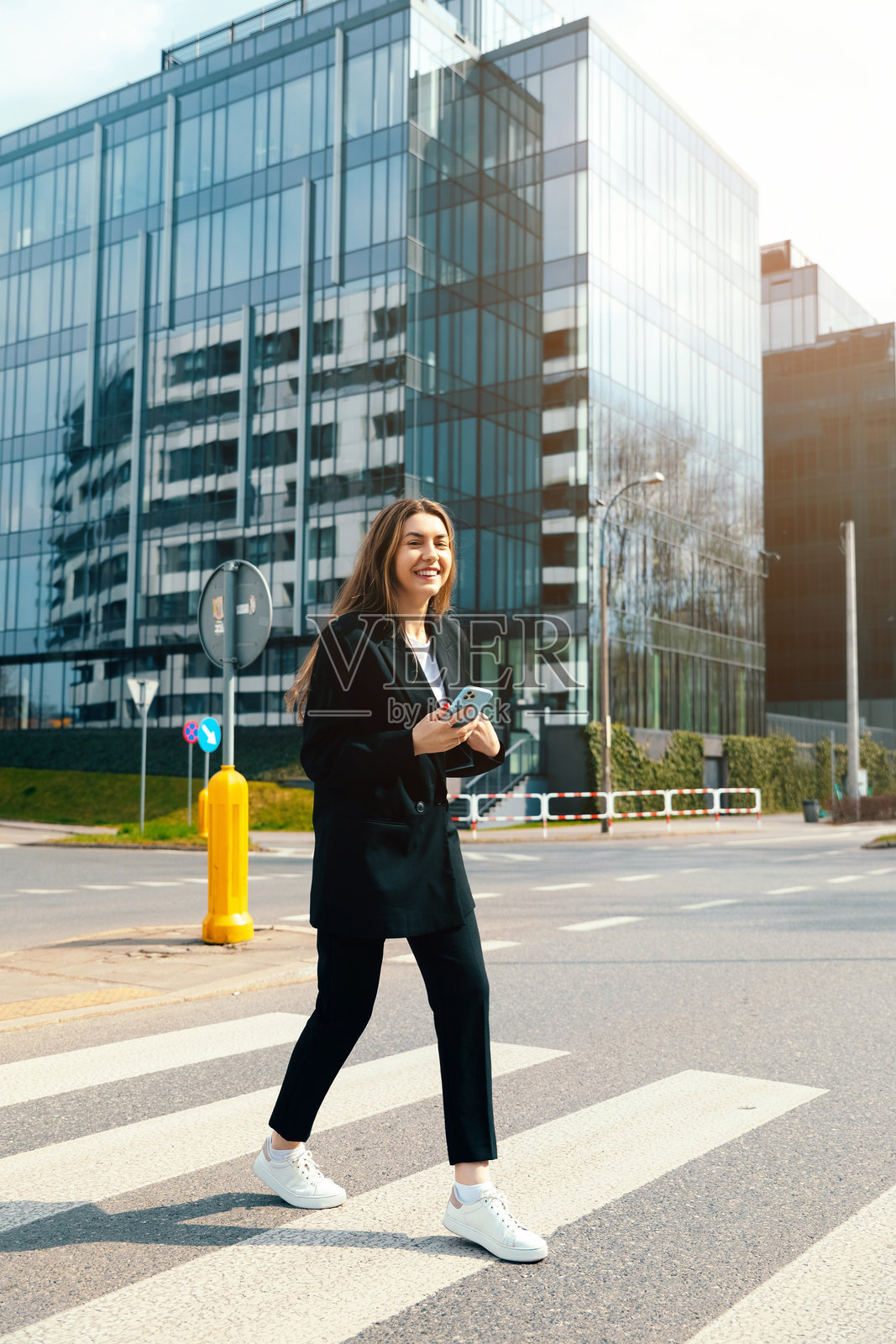自信的年轻女子手持智能手机走在城市街道上，身着时尚的黑色西装，背景是现代建筑，体现都市生活方式的概念照片摄影图片