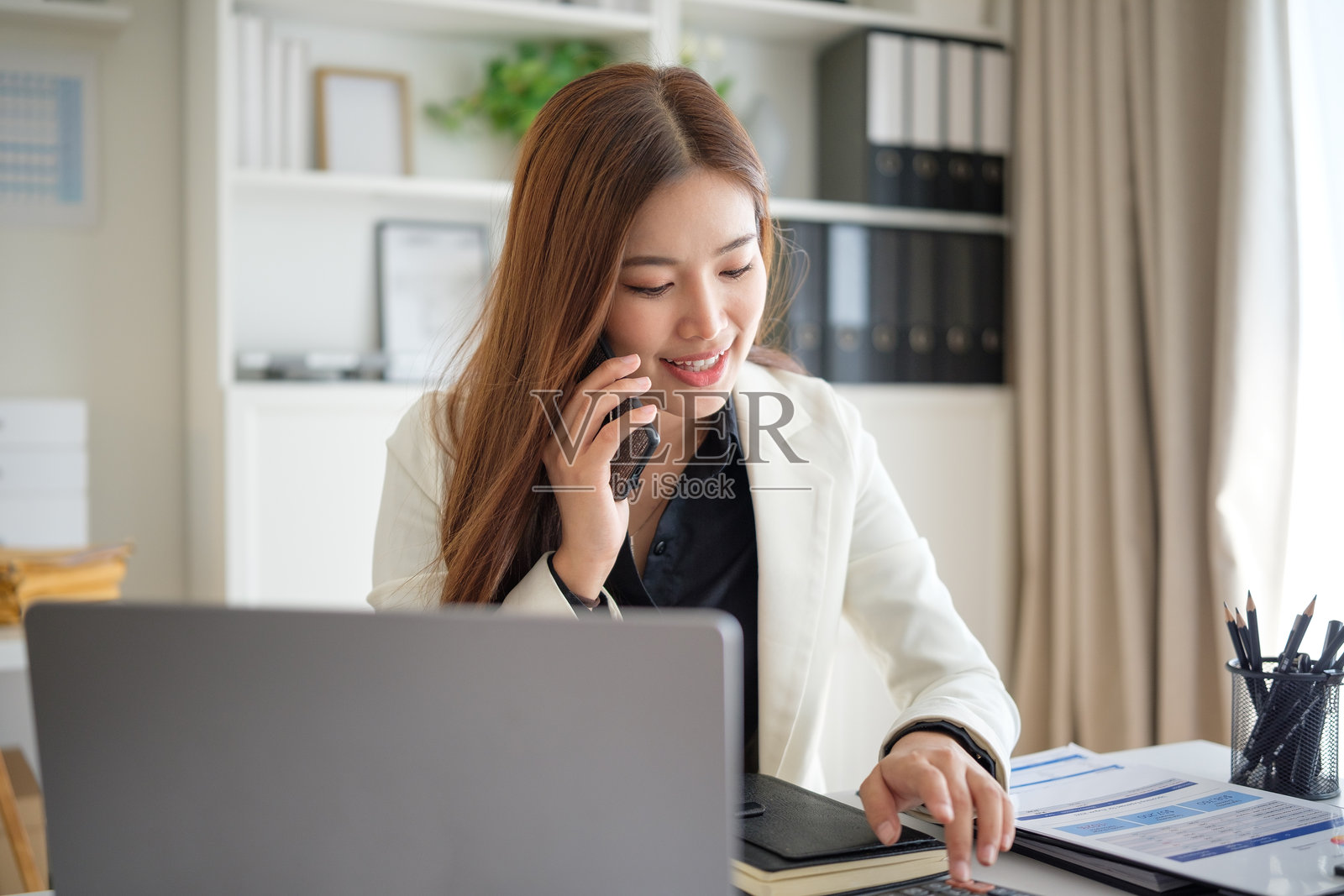 一位身穿白色西装外套的职业女性，在接听电话、谈论工作并指向文件时面带微笑，象征着商业规划与沟通。照片摄影图片