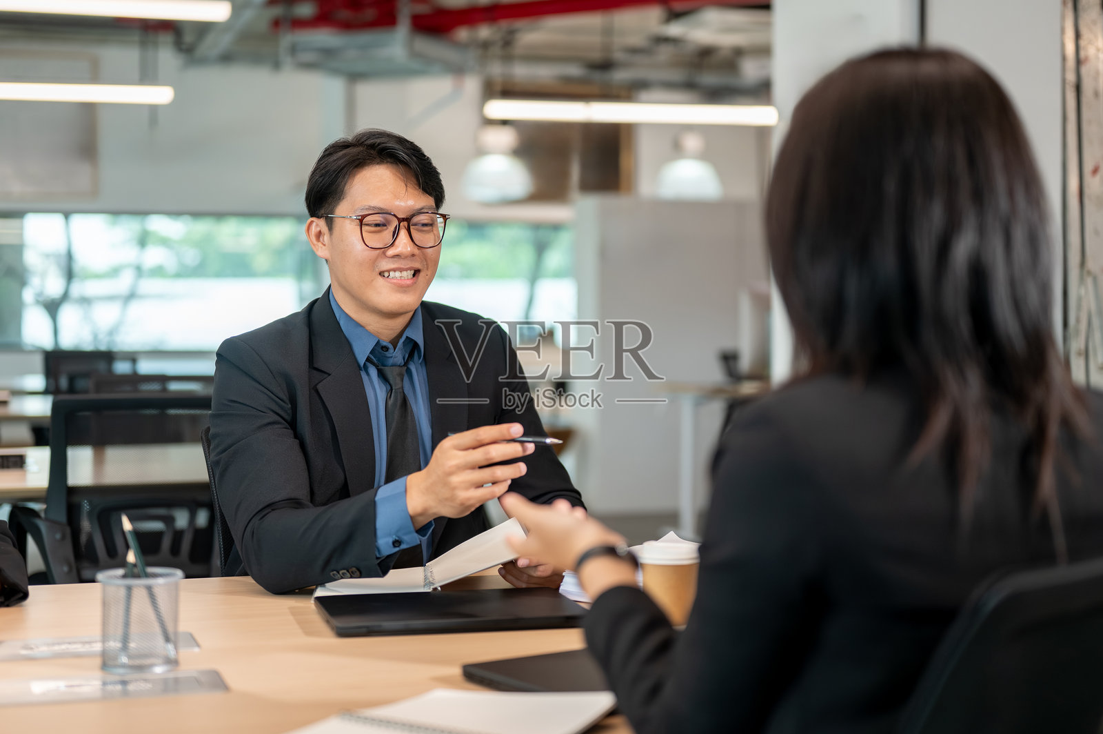 商务人士在会议室的木桌旁，与女性同事交谈，手持钢笔在笔记本上方照片摄影图片