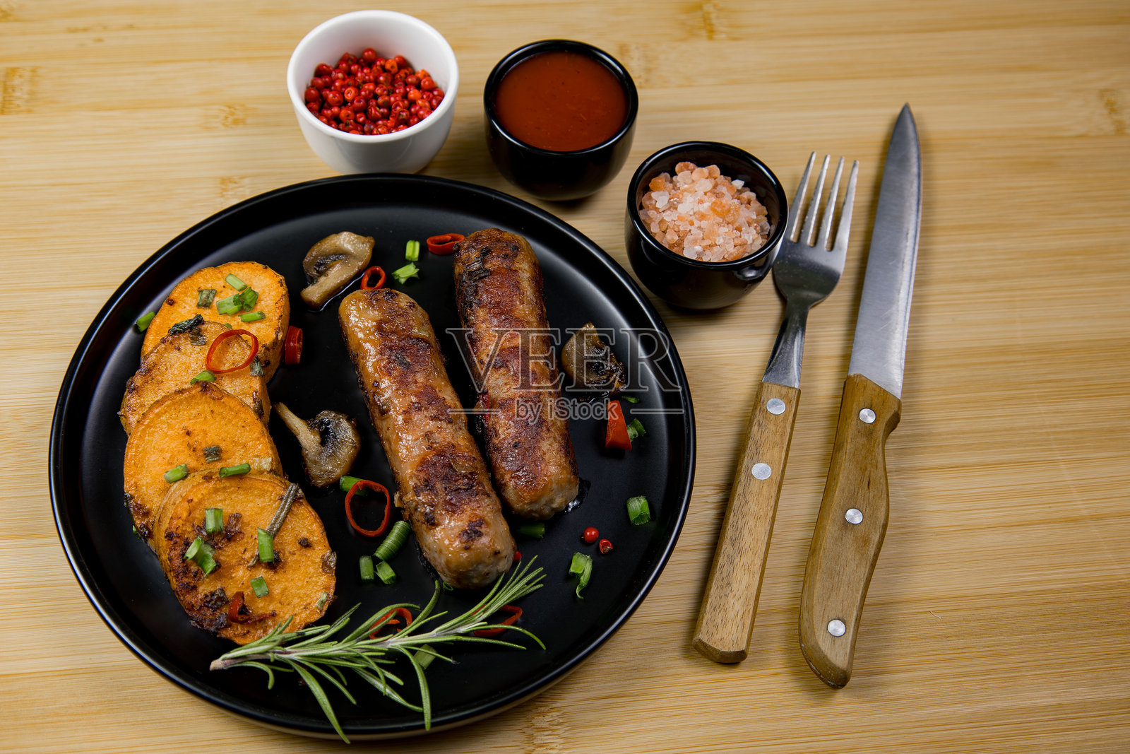 黑色盘子上放着美味的烤香肠和红薯片，配有蘸酱和餐具，摆放在木桌上照片摄影图片