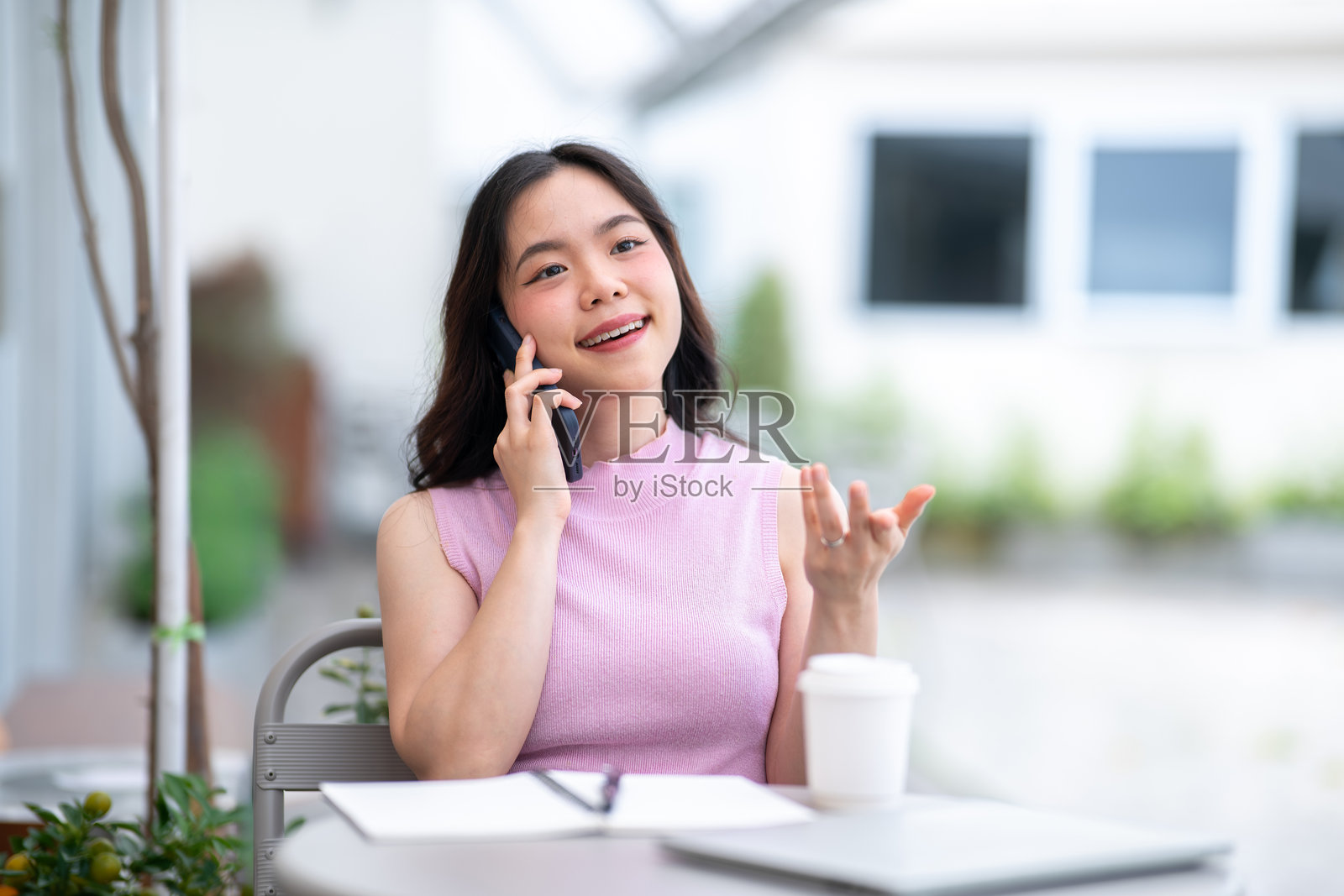 一位亚裔女学生在咖啡馆的桌子旁，一边看着笔记本电脑和书，一边用手比划着打电话交谈。照片摄影图片